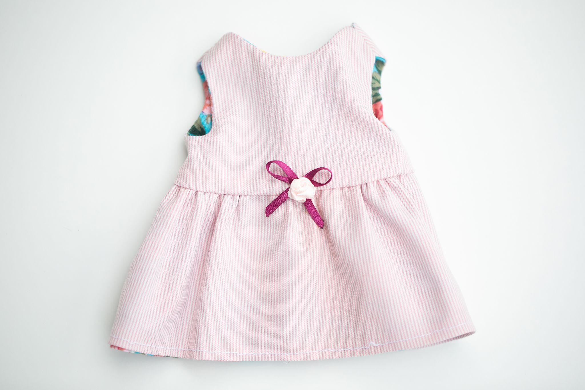 Pink sleeveless doll dress with floral trim, ribbon bow, and gathered skirt on white background.