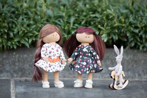 Two handmade cloth dolls with brown hair in floral dresses stand beside a small toy bunny on a rocking horse.