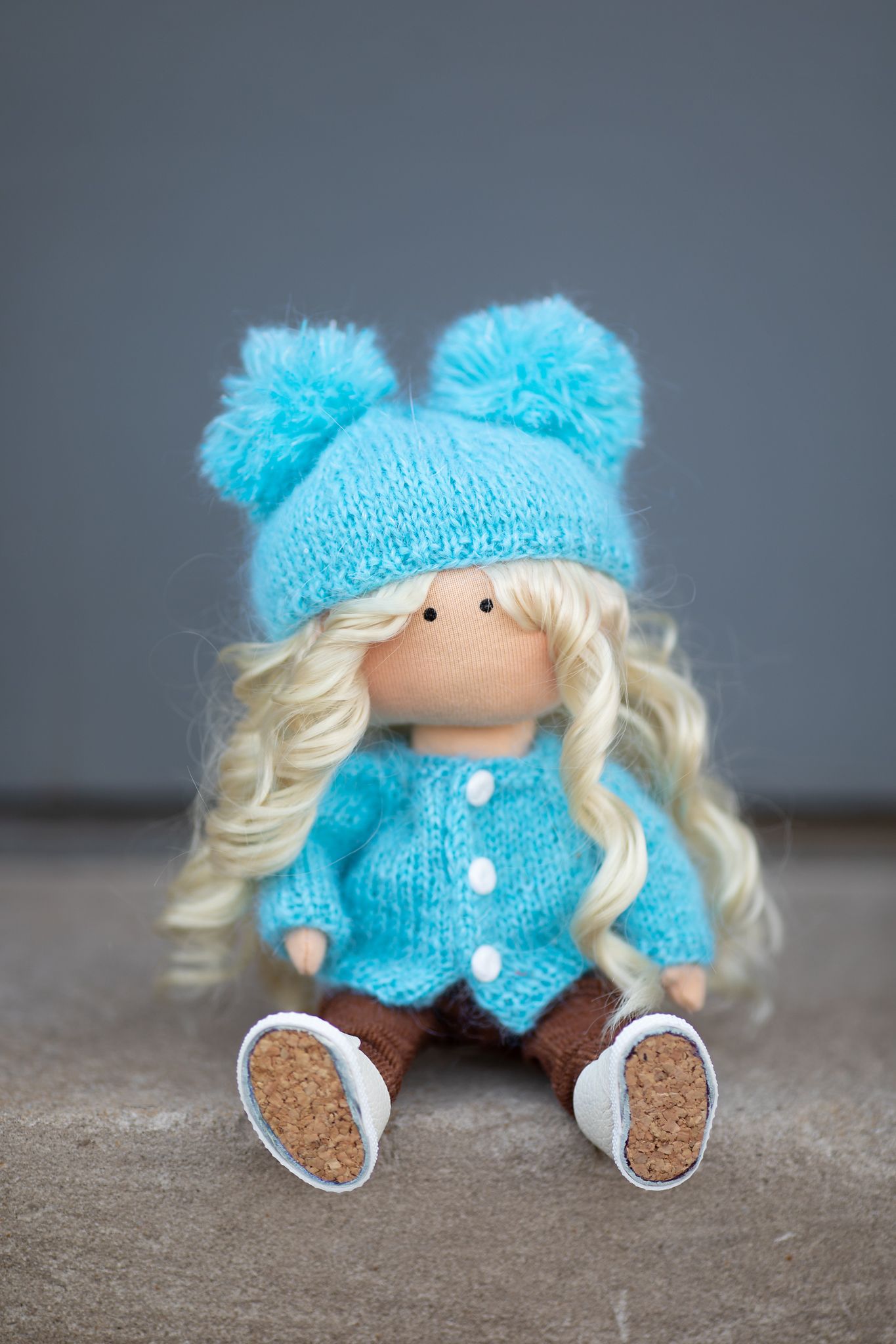 Handmade blonde doll wearing a turquoise knit hat with pompoms and matching blue cardigan sits on a surface.