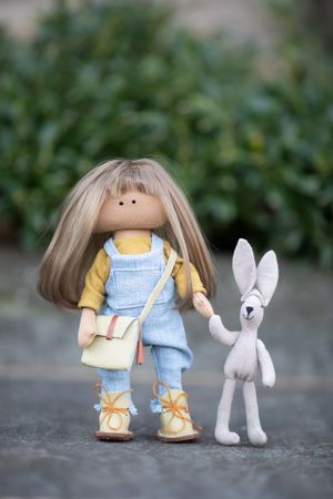 Handmade cloth doll with blonde hair in denim overalls holds hands with a small gray stuffed bunny toy.