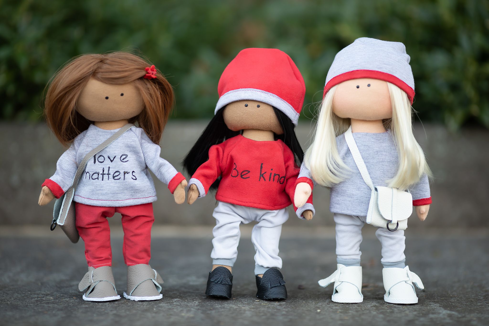 Three handmade dolls in red, white and gray casual outfits with inspirational message shirts stand together outdoors.