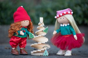 Two handmade Christmas elf dolls in red and green outfits stand beside a small wooden decorative holiday tree.