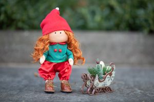 Handmade redhead elf doll in red and green outfit stands beside a miniature decorative sleigh with a tiny tree.