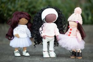 Three handmade dolls with dark hair in white and pink outfits stand together against a green leafy background.