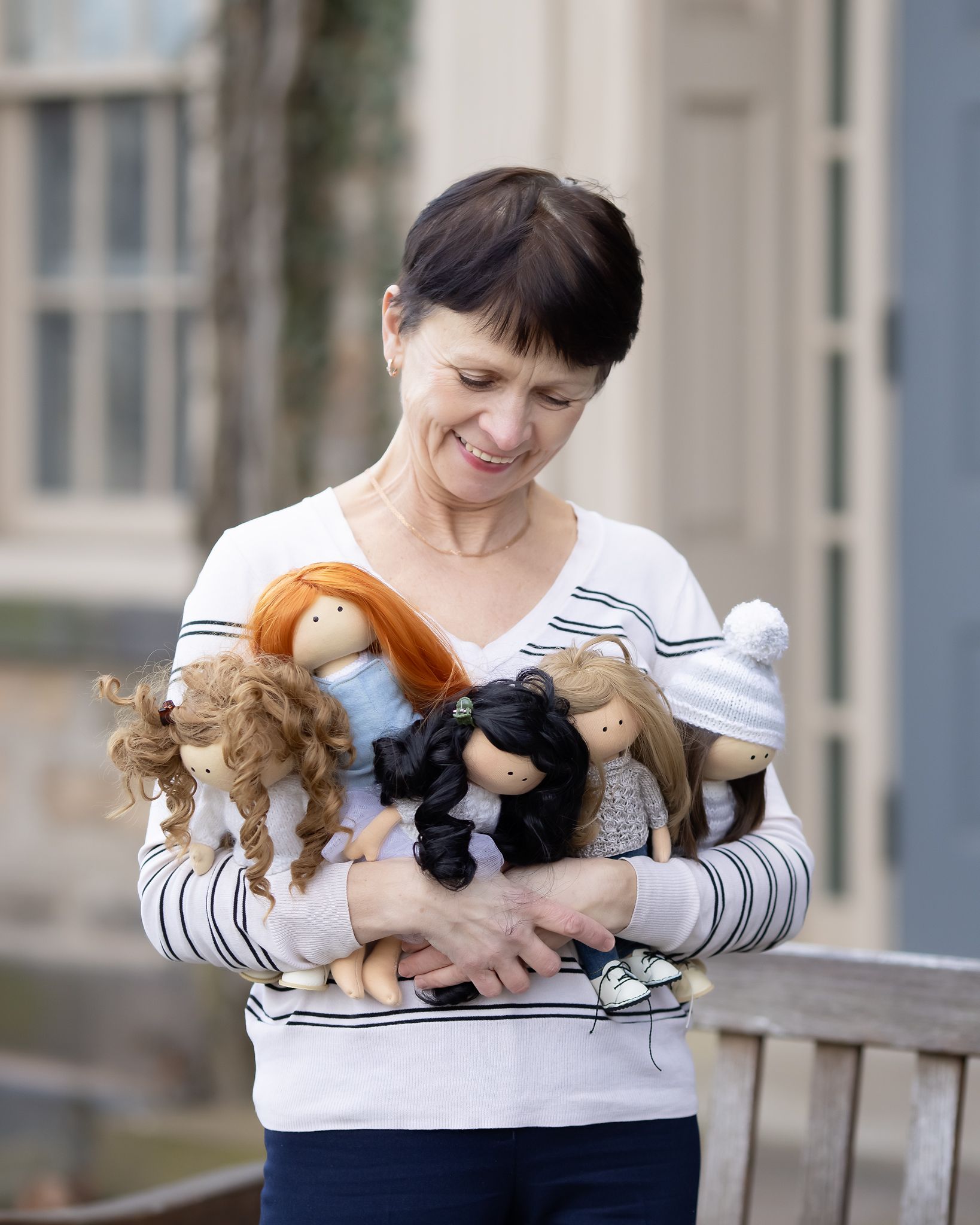 Smiling woman holding a collection of handmade fabric dolls with various hair colors outdoors.