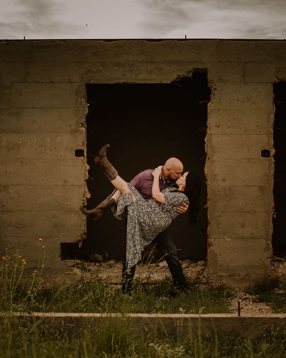 engaged couple kiss and dip while kicking her boots up wearing a flower dress wrapping her arms around his neck