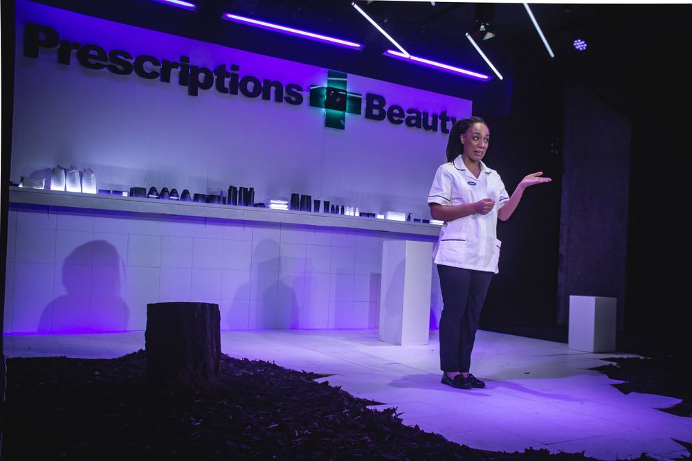 A performer gestures while standing near a pharmacy counter set with purple theatrical lighting.
