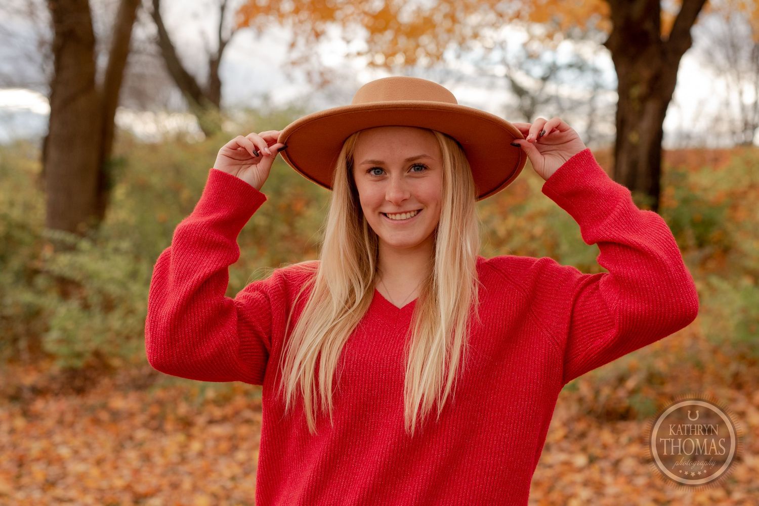 Playful autumn portrait in red sweater holding wide brim hat among fall foliage.