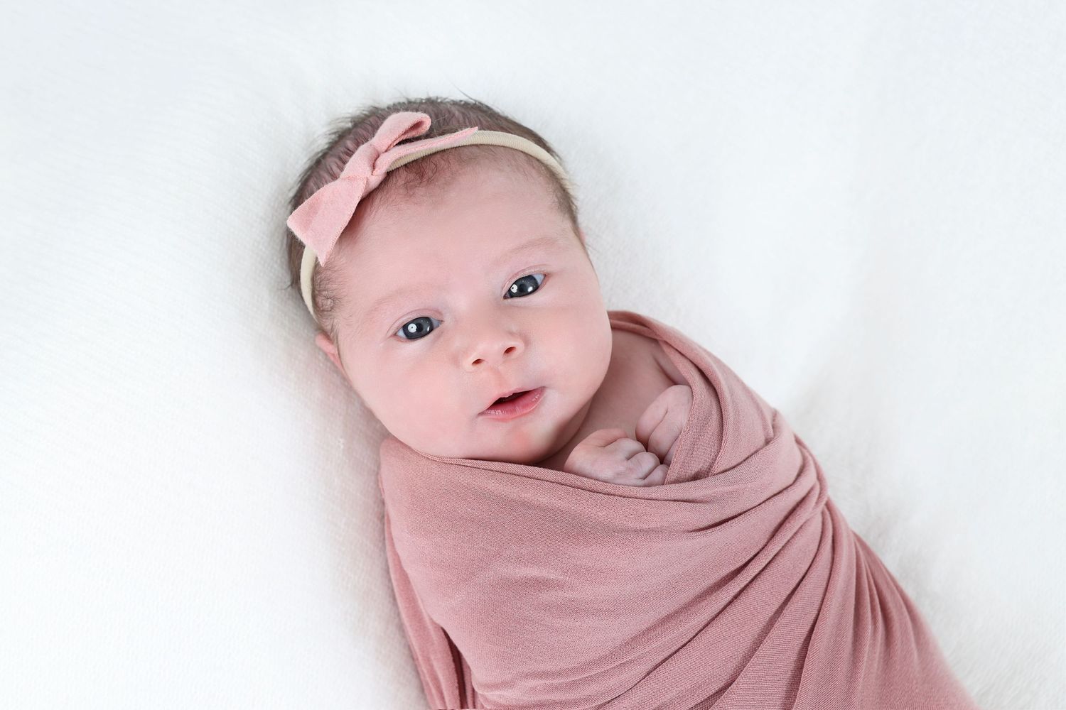 Alert newborn wrapped in a soft pink swaddle with matching bow . her hands are peeking out of the blanket.