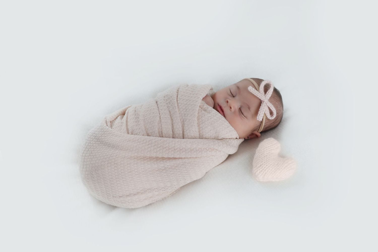 Sleeping newborn wrapped in a soft white swaddle with a matching knit heart nearby
