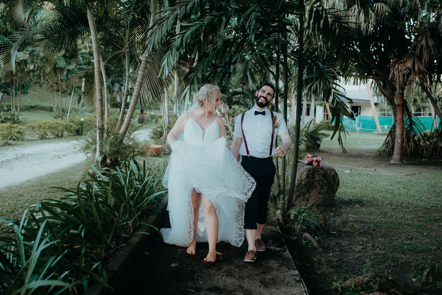 A couple in wedding attire walking together on a tropical garden path surrounded by palm trees and lush greenery.