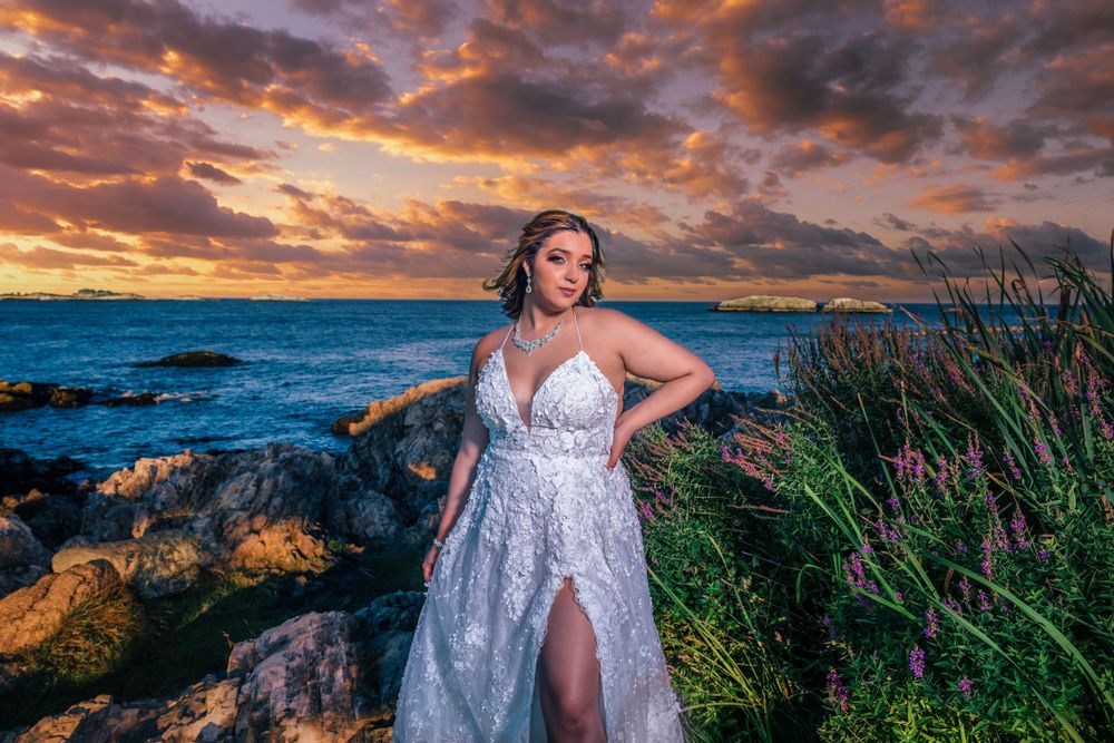 A dreamy coastal photoshoot features a flowing white dress against vibrant sunset clouds.