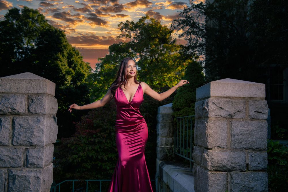 Figure in deep pink formal dress poses between stone pillars at dusk.