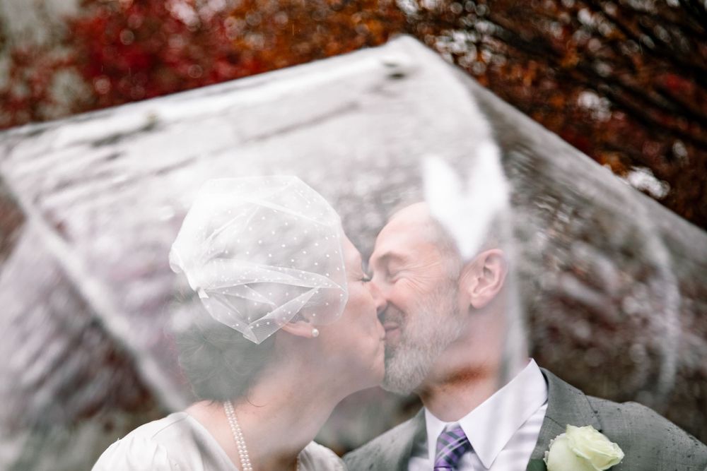 A romantic wedding photo of a couple kissing under a veil with fall foliage in the background.