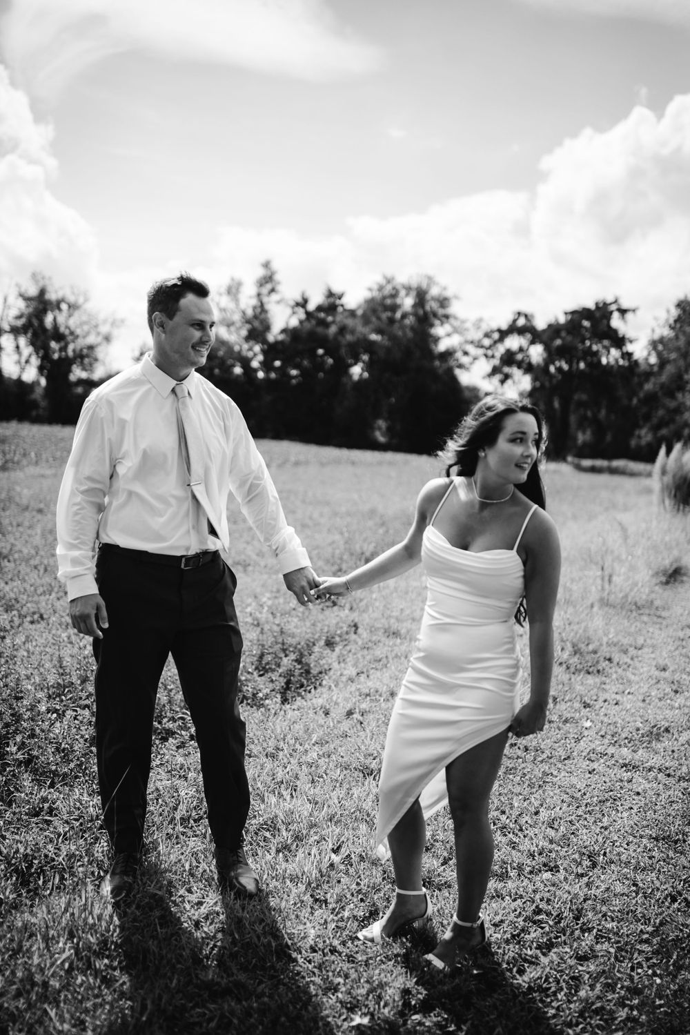 A black and white photo of a couple walking through a meadow holding hands.