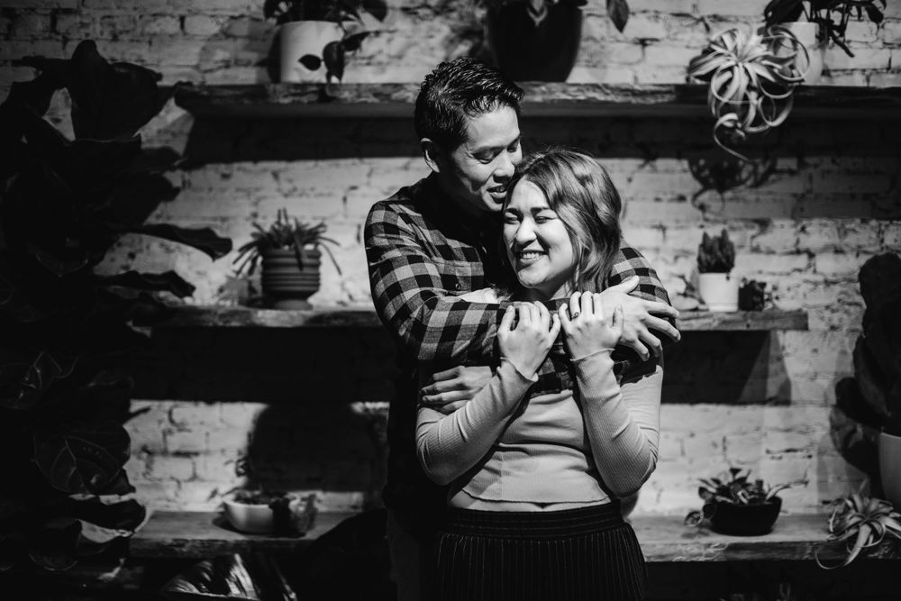 A black and white photo of a couple embracing against a brick wall with plants.