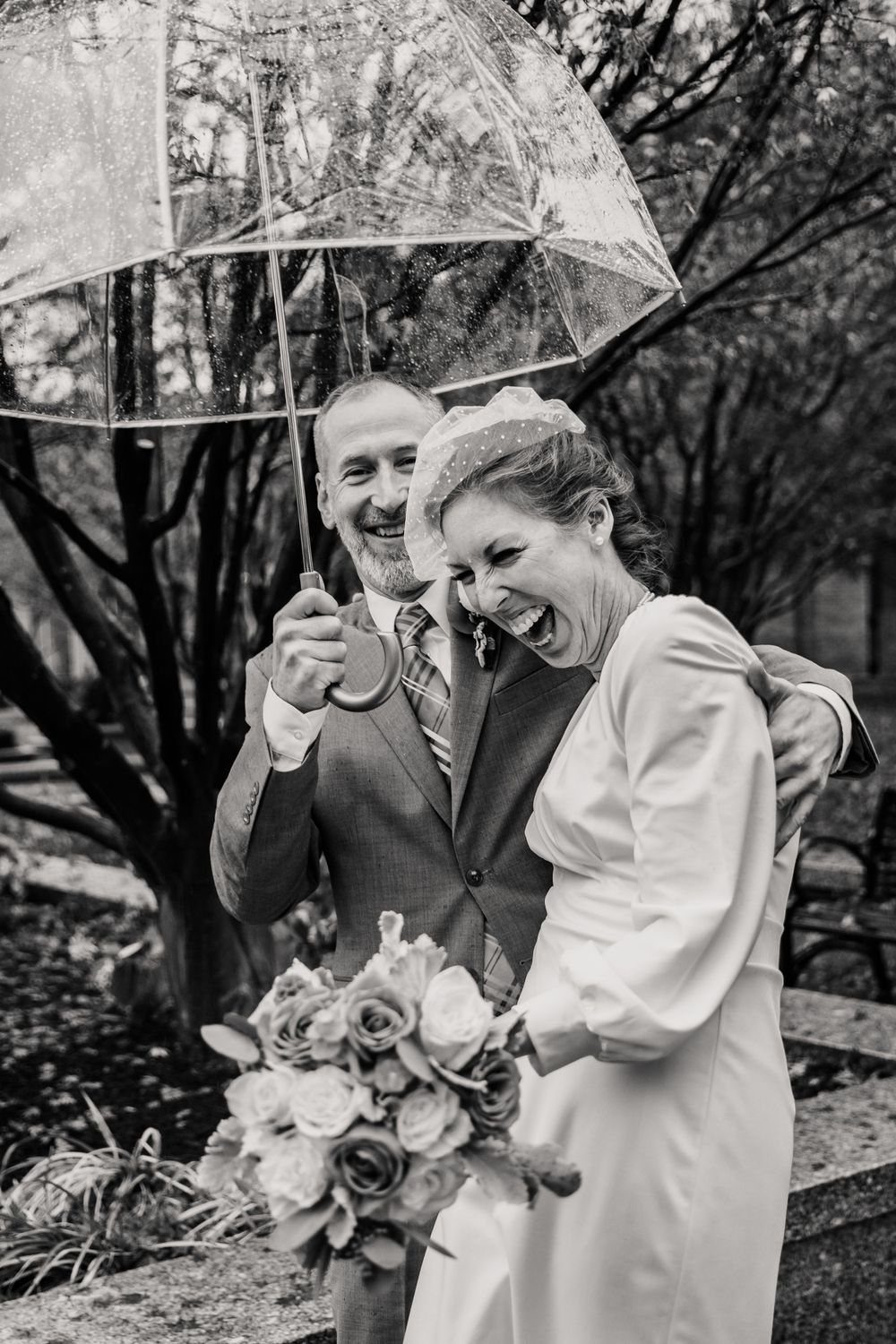 A couple sharing a joyful moment under a clear umbrella in the rain on their wedding day.