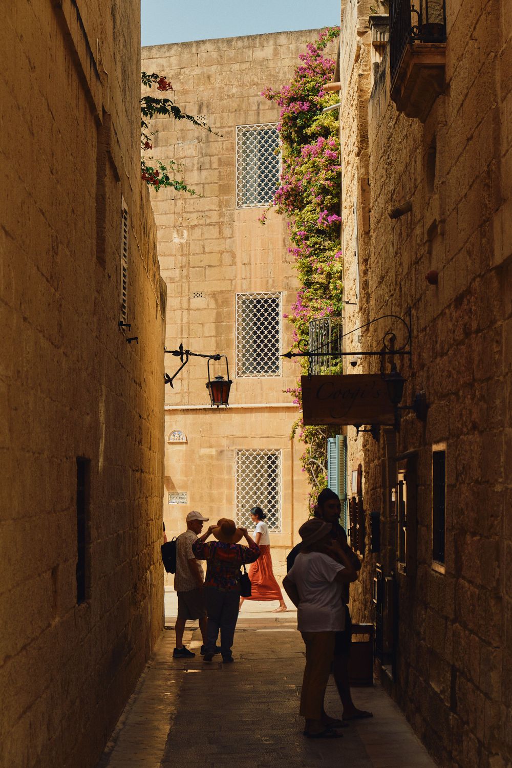 Tourists walk through a narrow stone alley with latticed windows and pink bougainvillea flowers in Malta.
