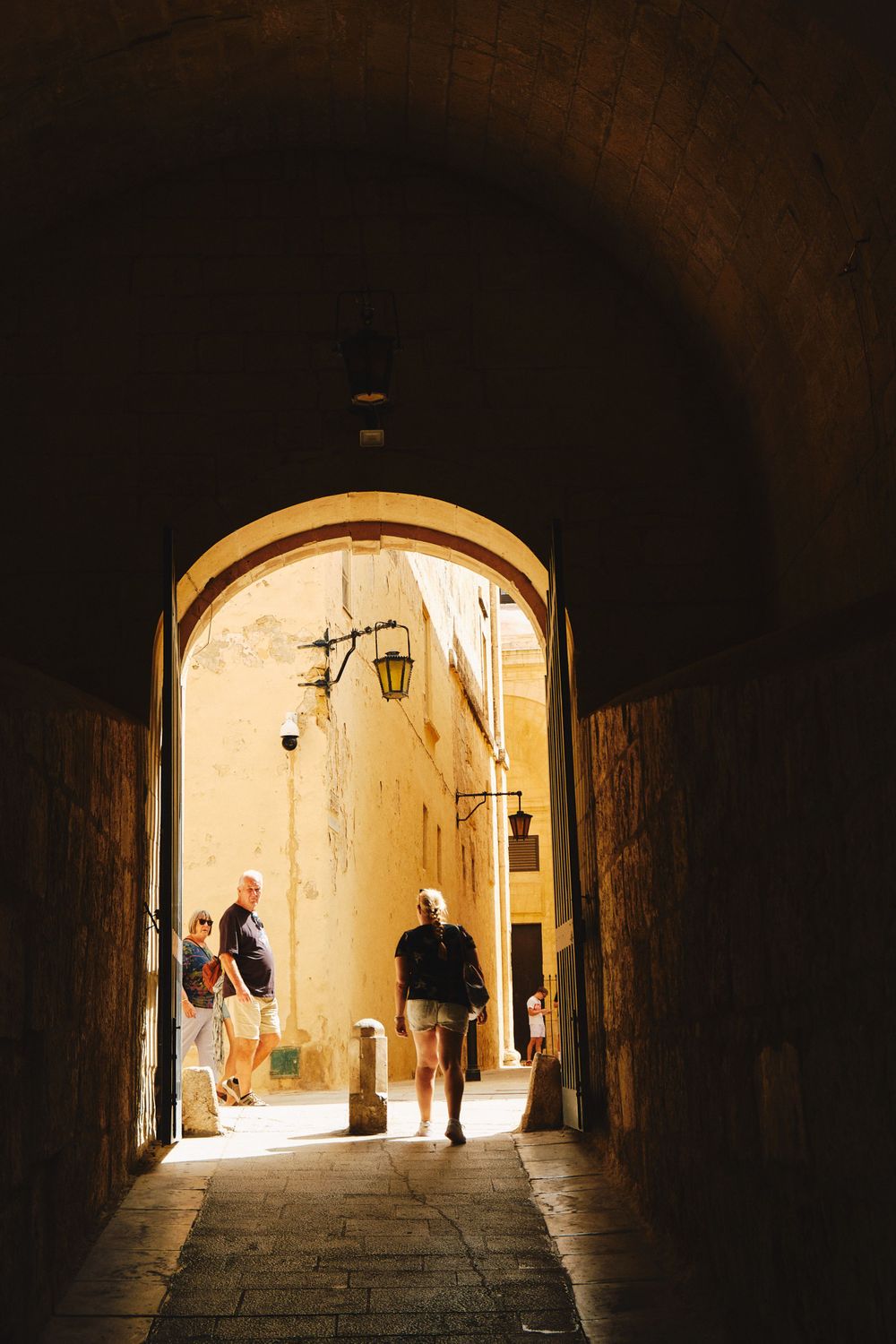 Sunlit archway passage in ancient Mediterranean stone building with tourists walking through.