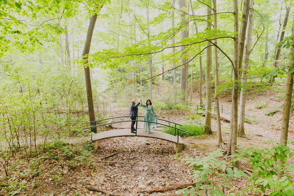 A couple stands together on a wooden footbridge in a serene forest setting.