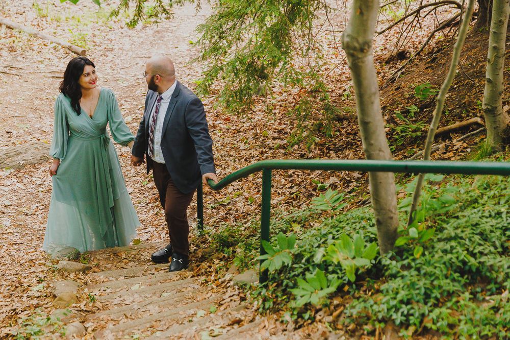 A couple walks along a wooded path with fallen leaves, the woman wearing a flowing mint green dress.