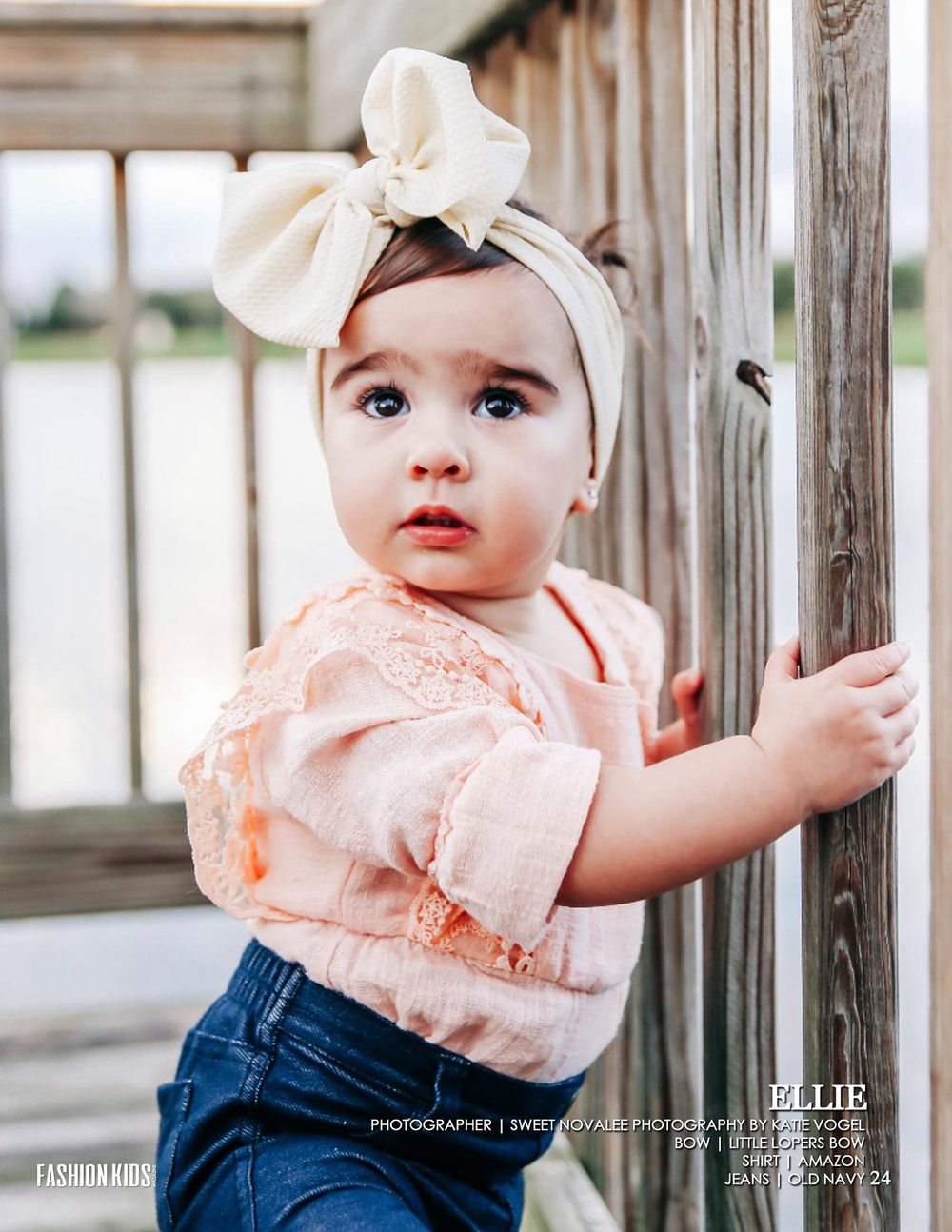 Young toddler in pink blouse and denim with cream bow headband standing by wooden railing.
