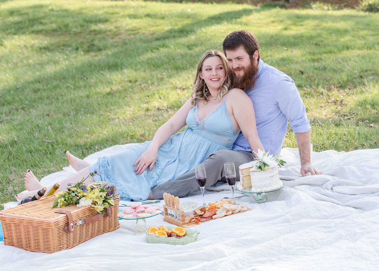 A romantic picnic setup on a white blanket in a grassy park with a wicker basket and snacks.
