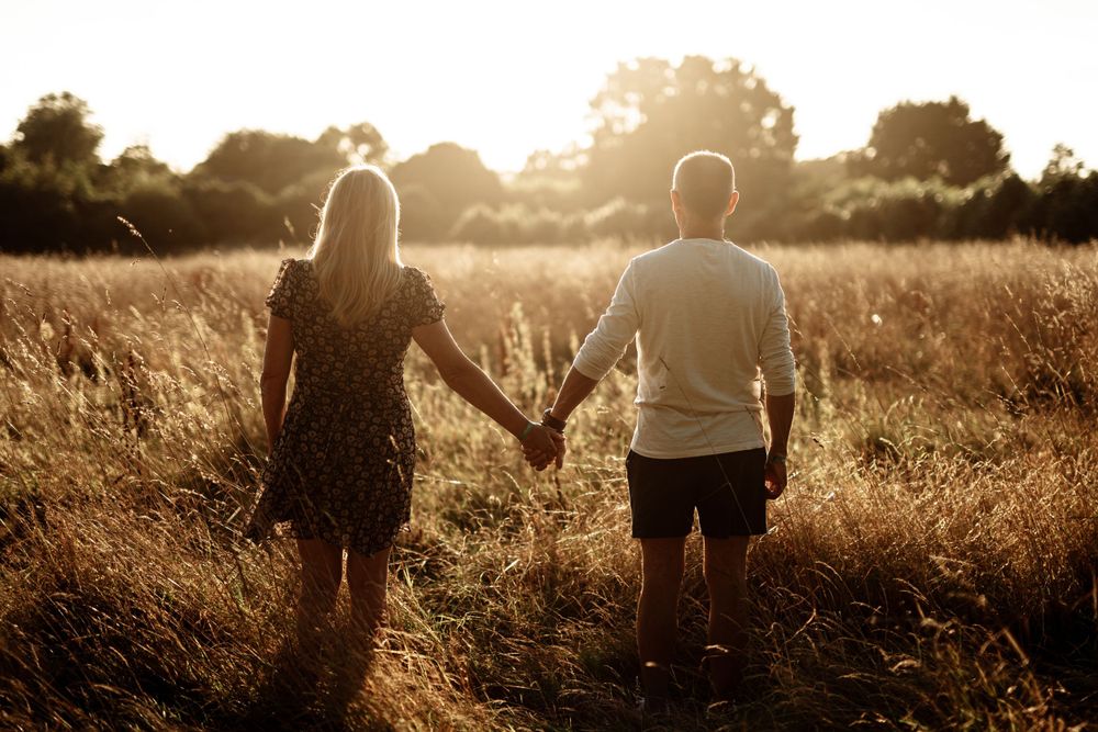 Backlit couple holding hands while walking through golden wheat field at sunset.