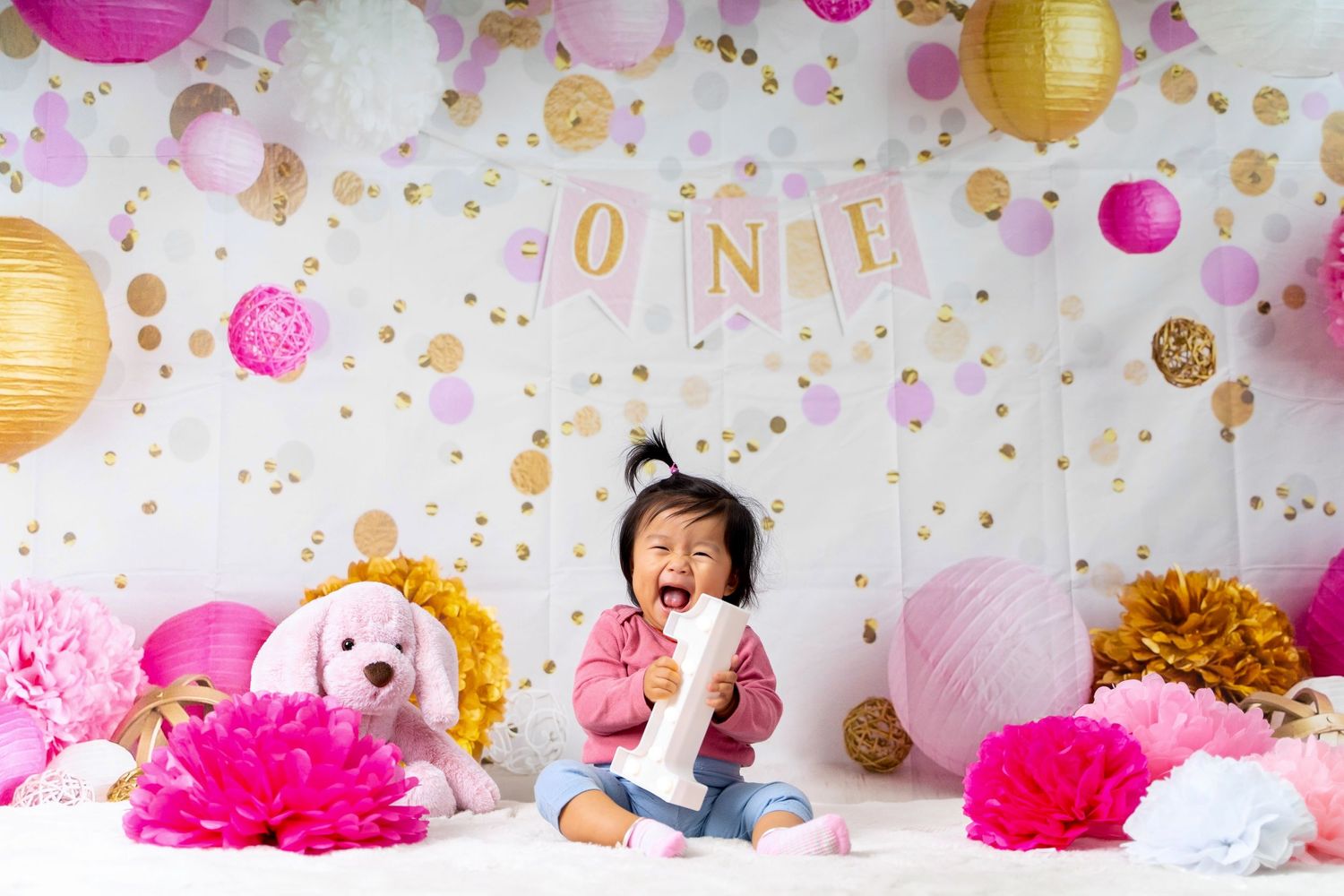 Colorful first birthday party setup with pink and gold decorations, lanterns, and plush toys.