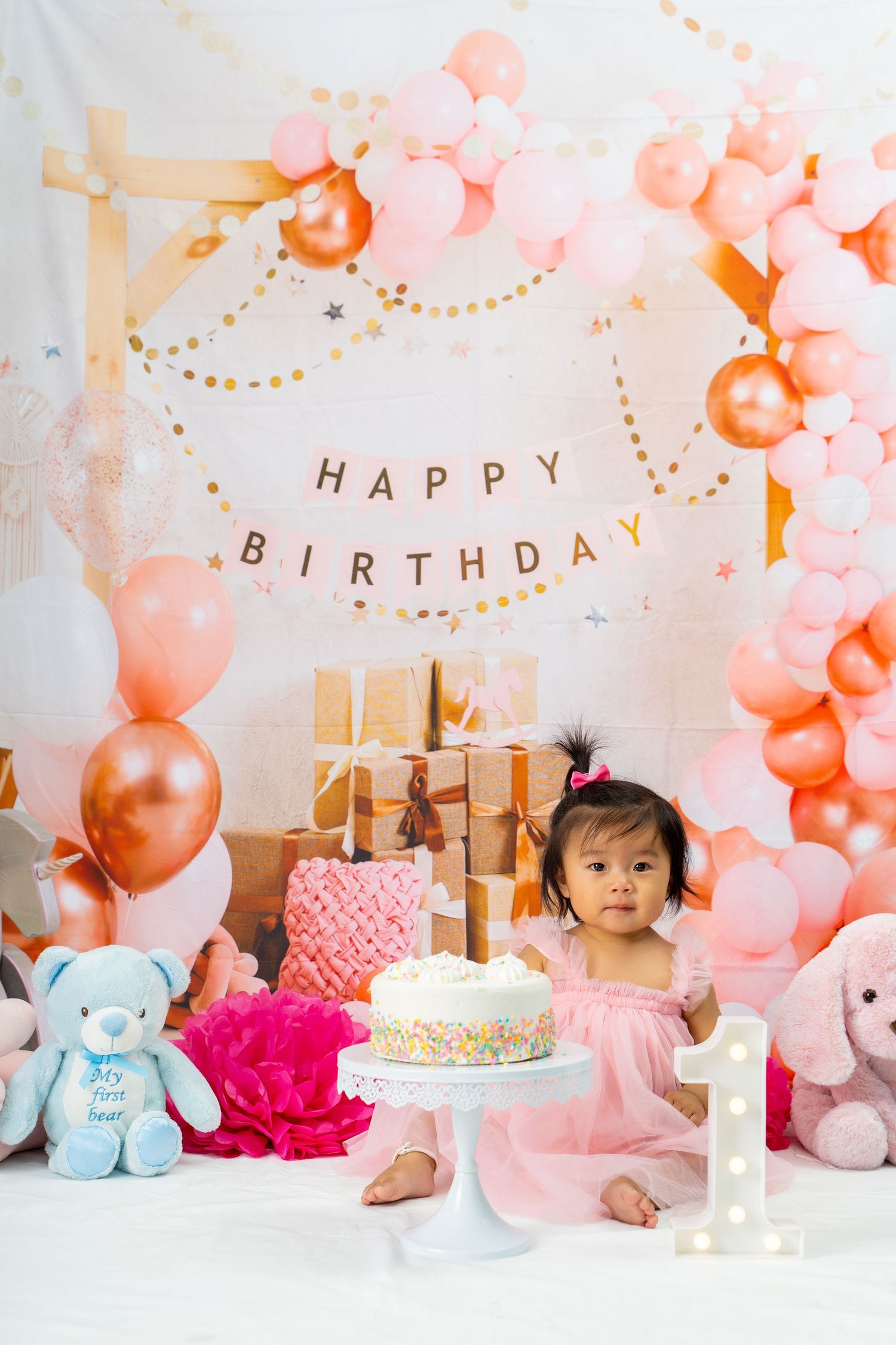 Happy Birthday decoration with pink and rose gold balloons, cake, and presents against white backdrop.