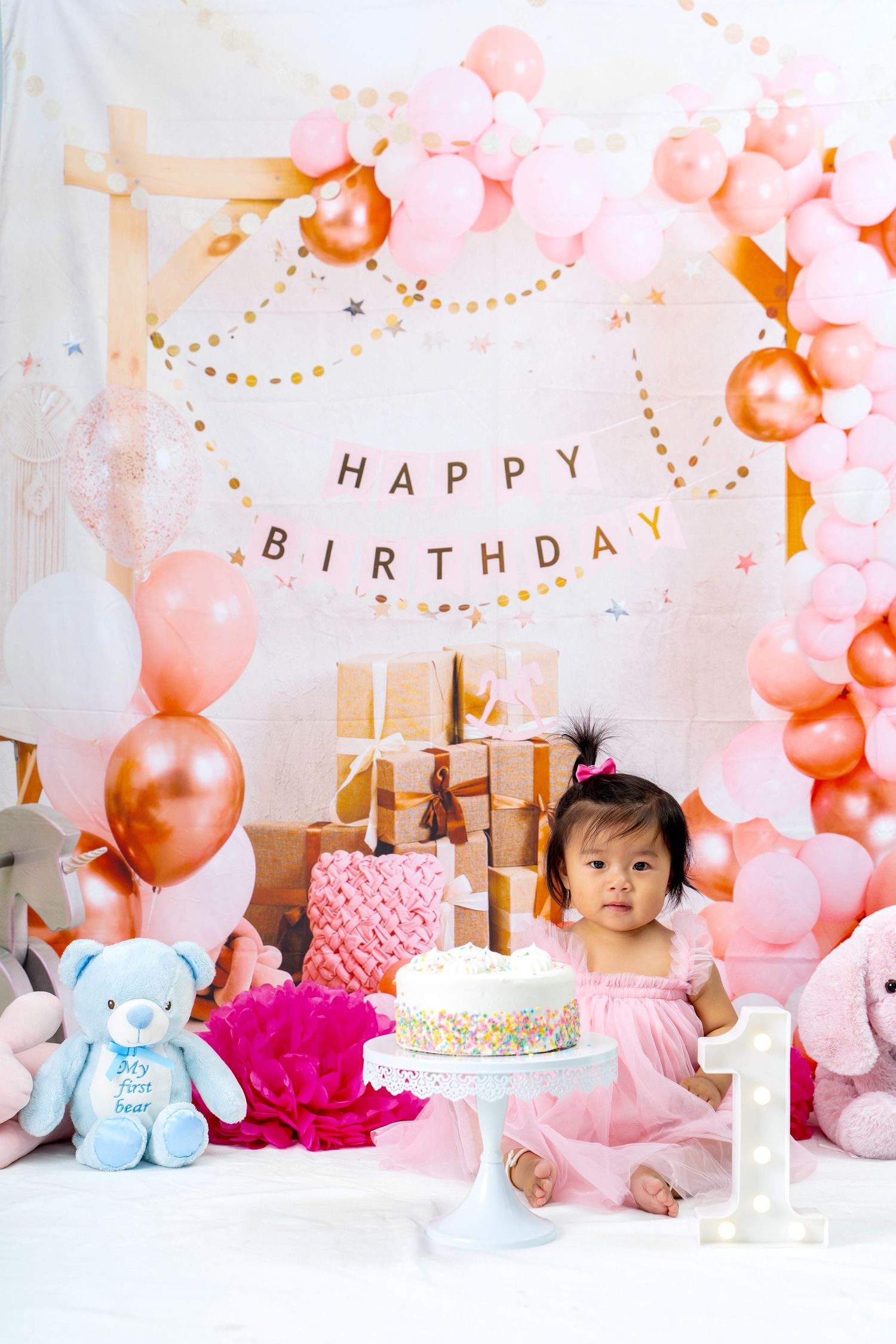 First birthday party setup with pink balloons, birthday cake, stuffed animals and wrapped presents.