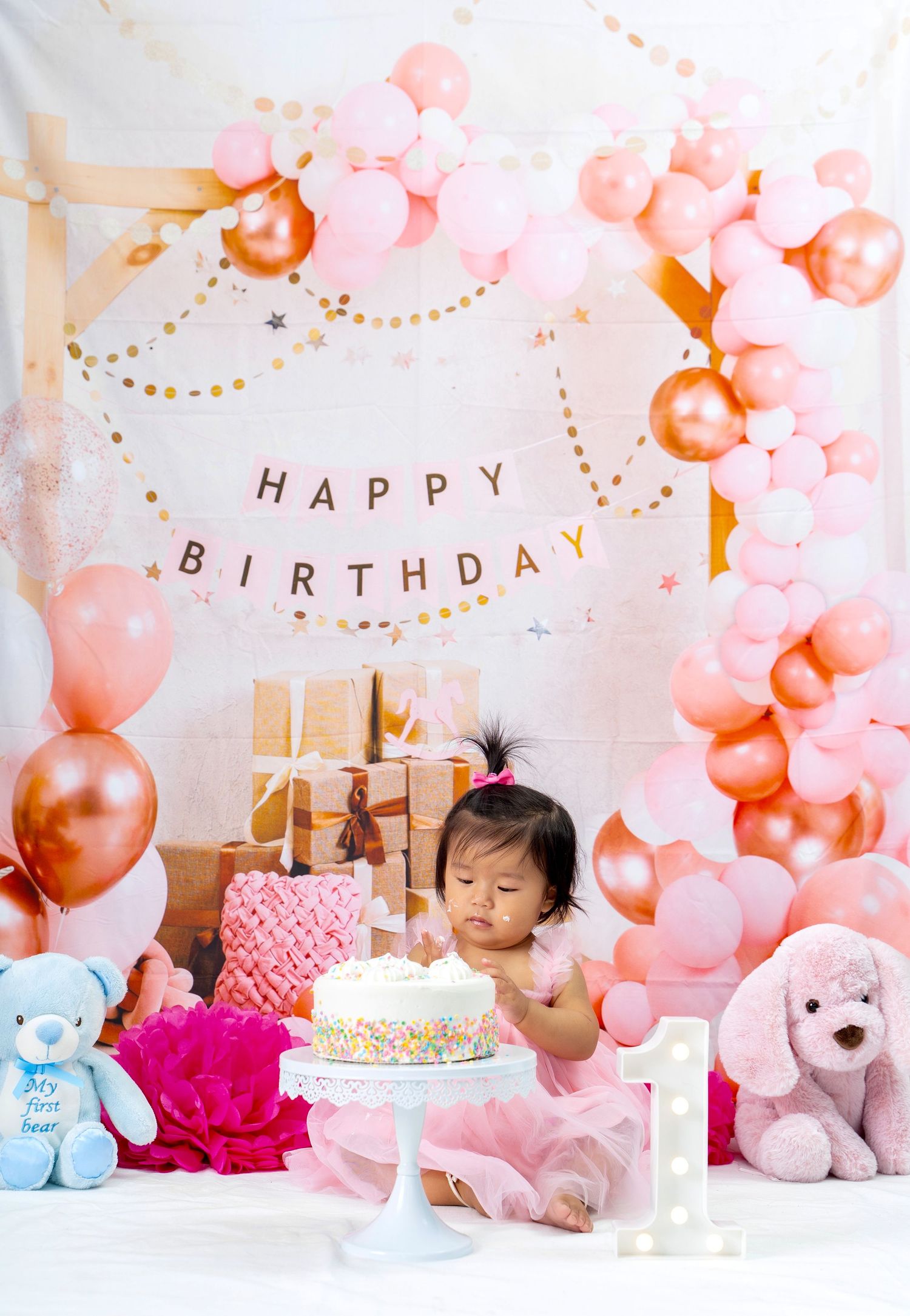 Pink and gold birthday party scene with cake, presents, and plush toys against decorated backdrop.