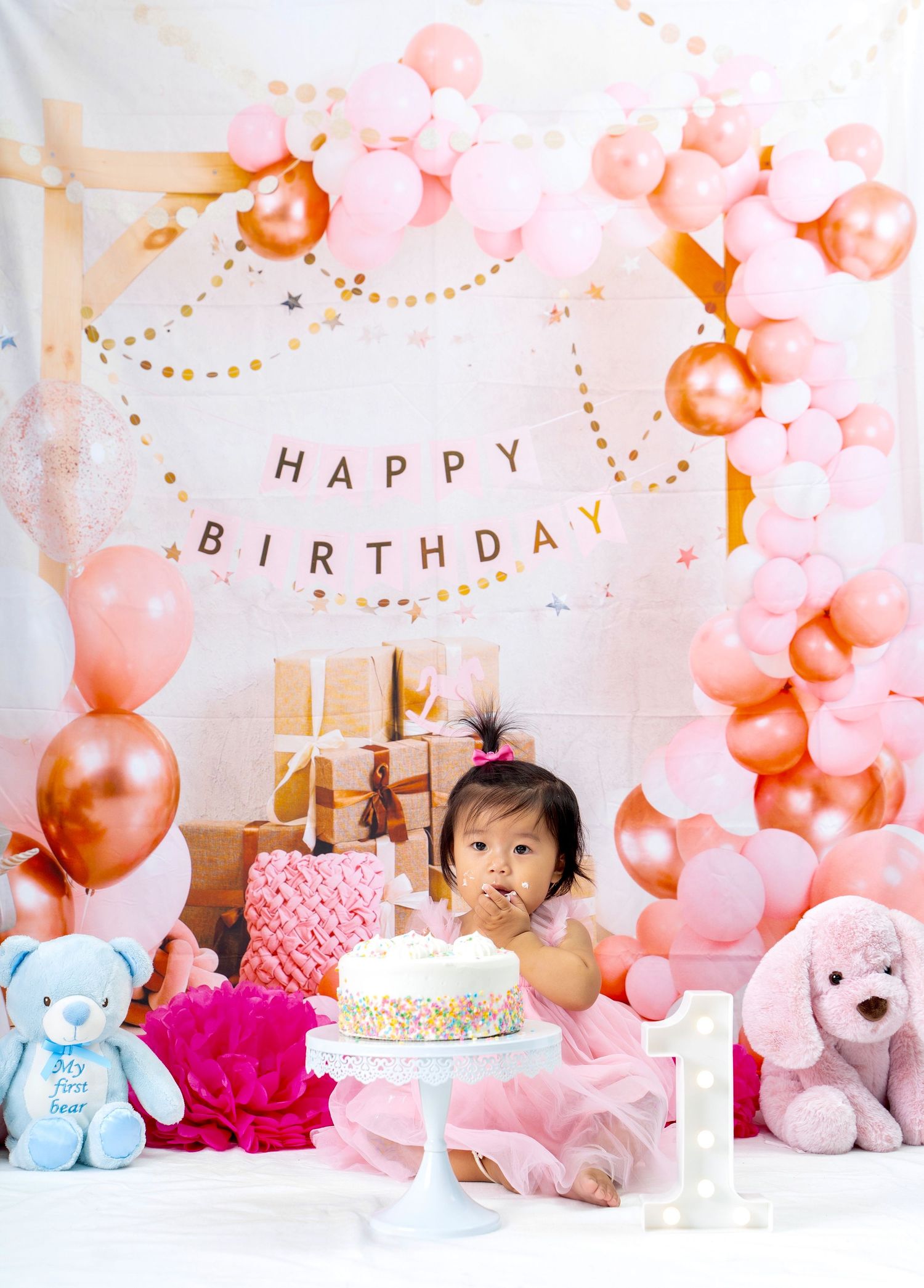 First birthday celebration with pink balloons, white cake, and stuffed animals on decorated background.
