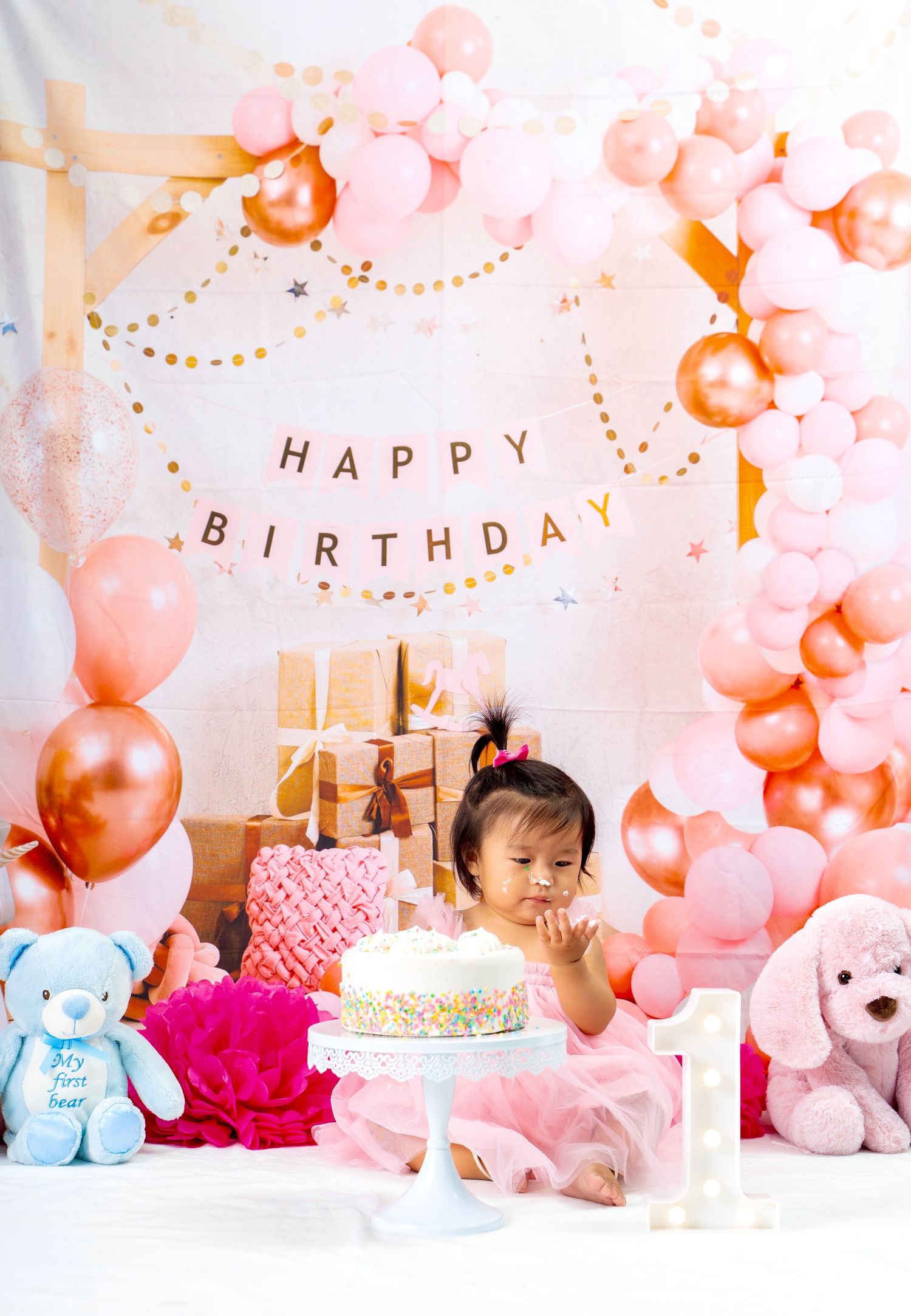 Festive first birthday setup with balloon arch, wrapped gifts, and birthday cake on white backdrop.