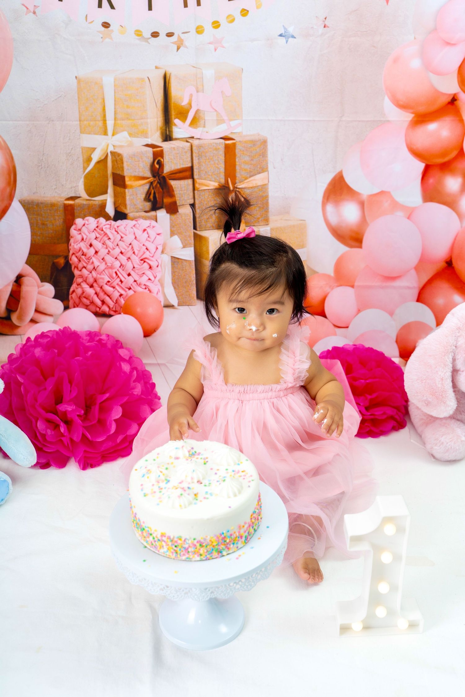Pink themed first birthday party with decorative balloons, wrapped presents and sprinkle-covered cake.