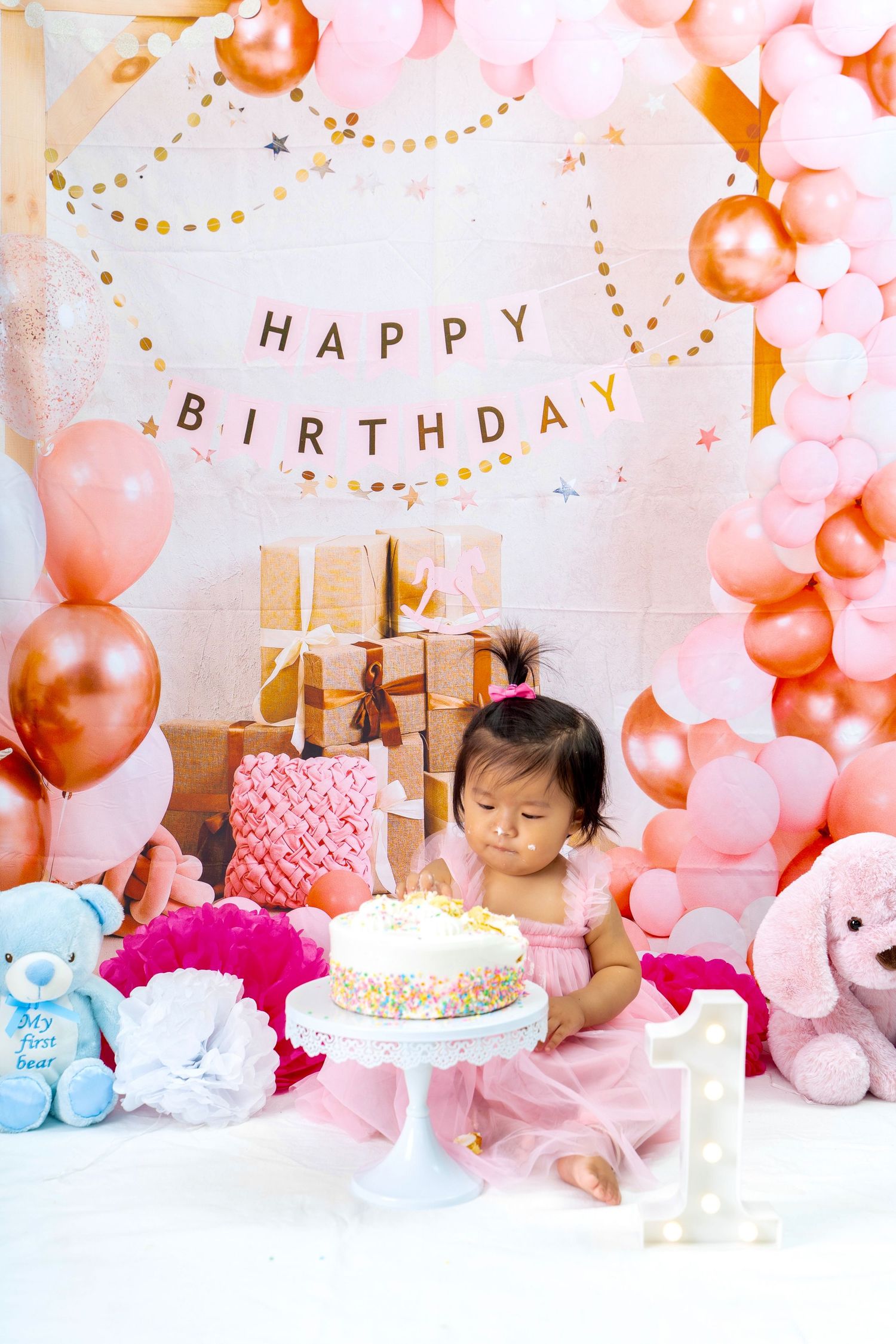 First birthday celebration with pink and gold balloon decorations, plush toys and white frosted cake.