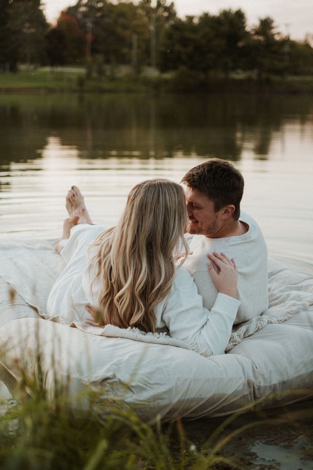 Couple in white lounging on blanket by serene lake at sunset.
