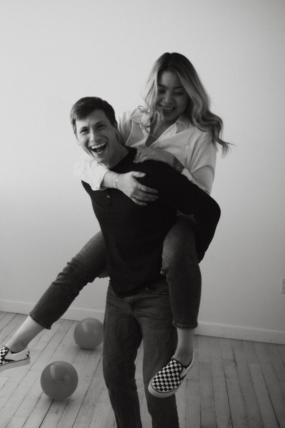 A playful black and white photo of a piggyback ride moment with a couple in casual attire on a wooden floor.
