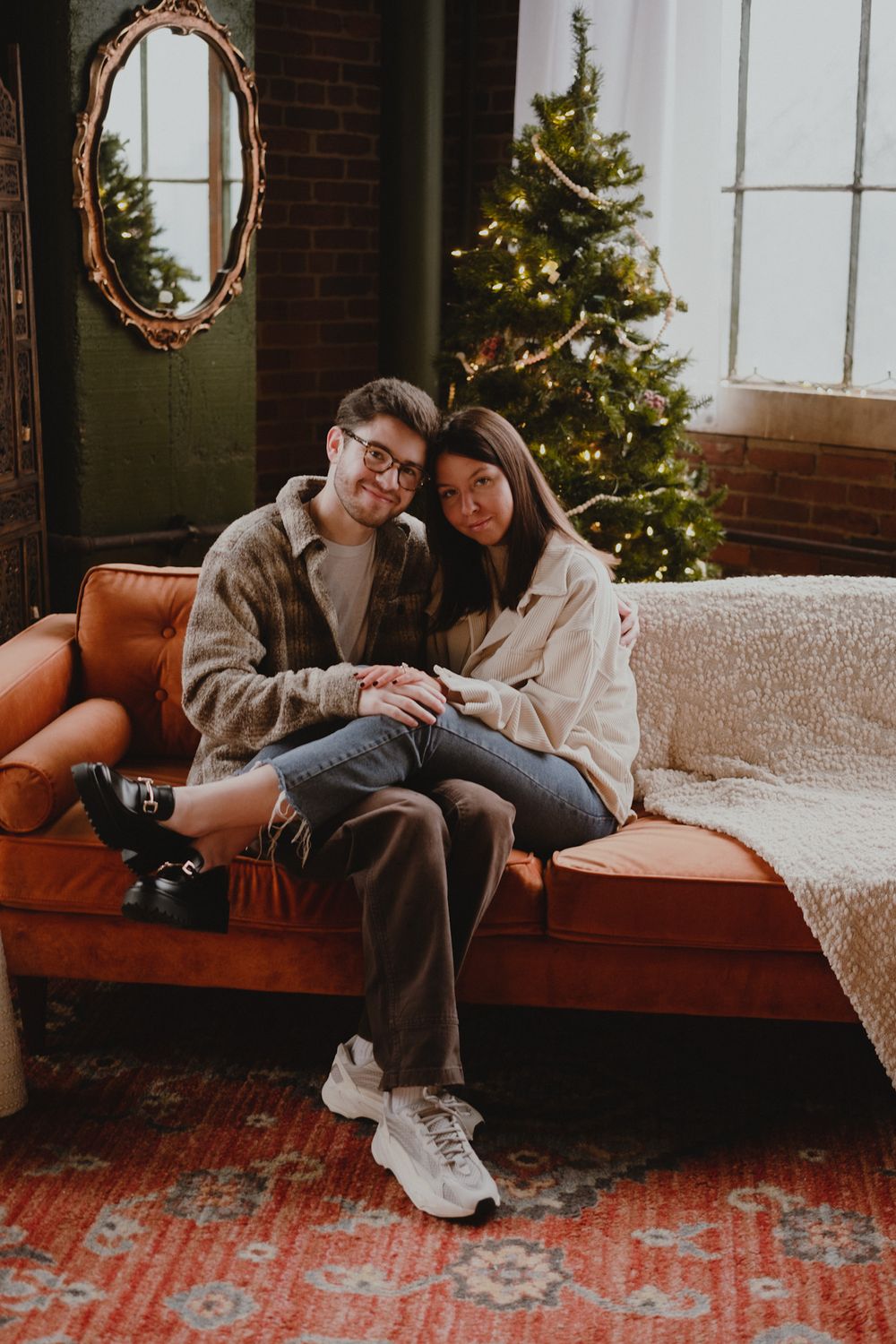 Cozy Christmas scene with a couple lounging on a sofa near a decorated tree and brick wall.