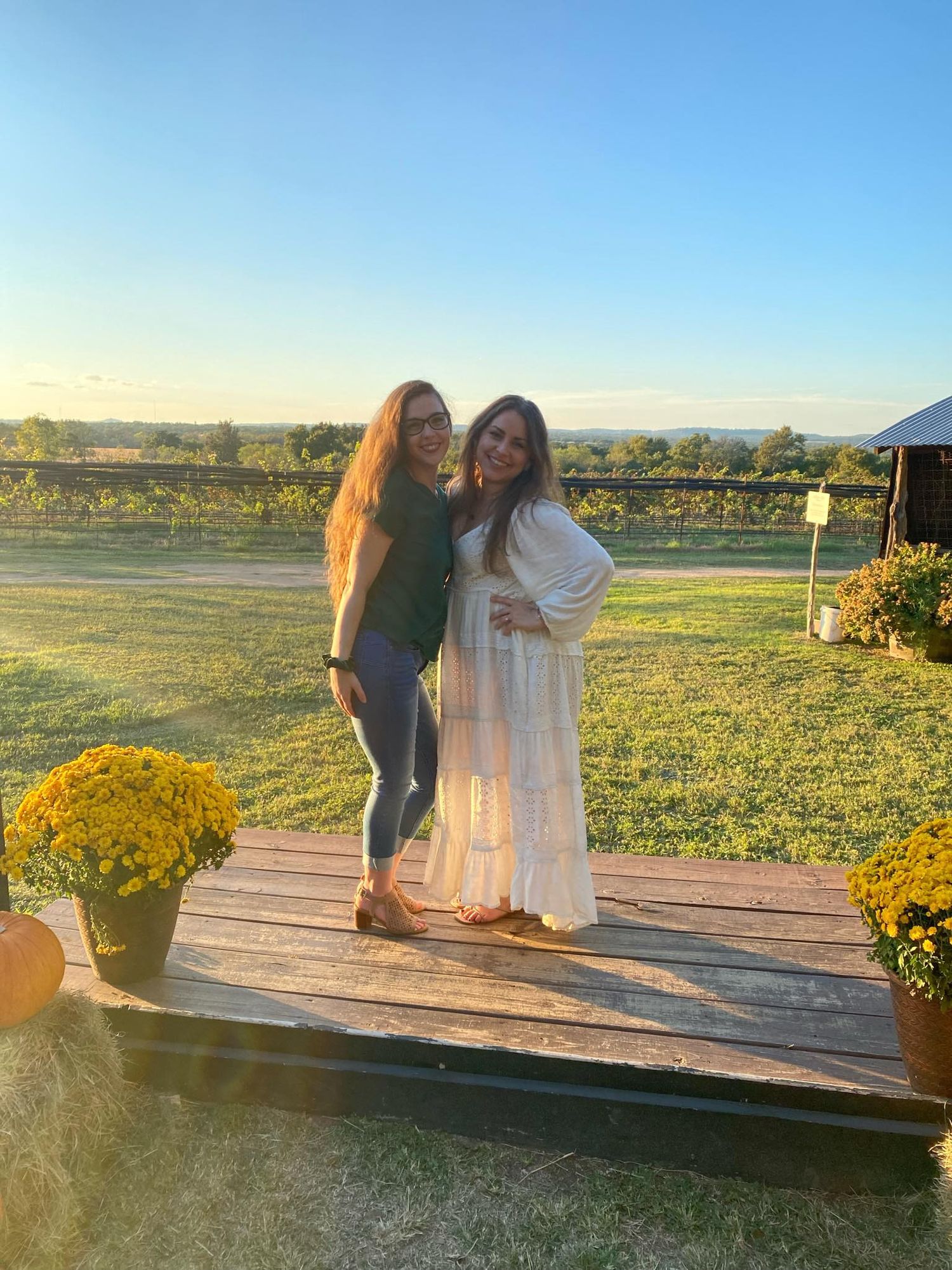 A long haired brunette woman wearing green poses with her brunette friend wearing a white dress with grape vines behind