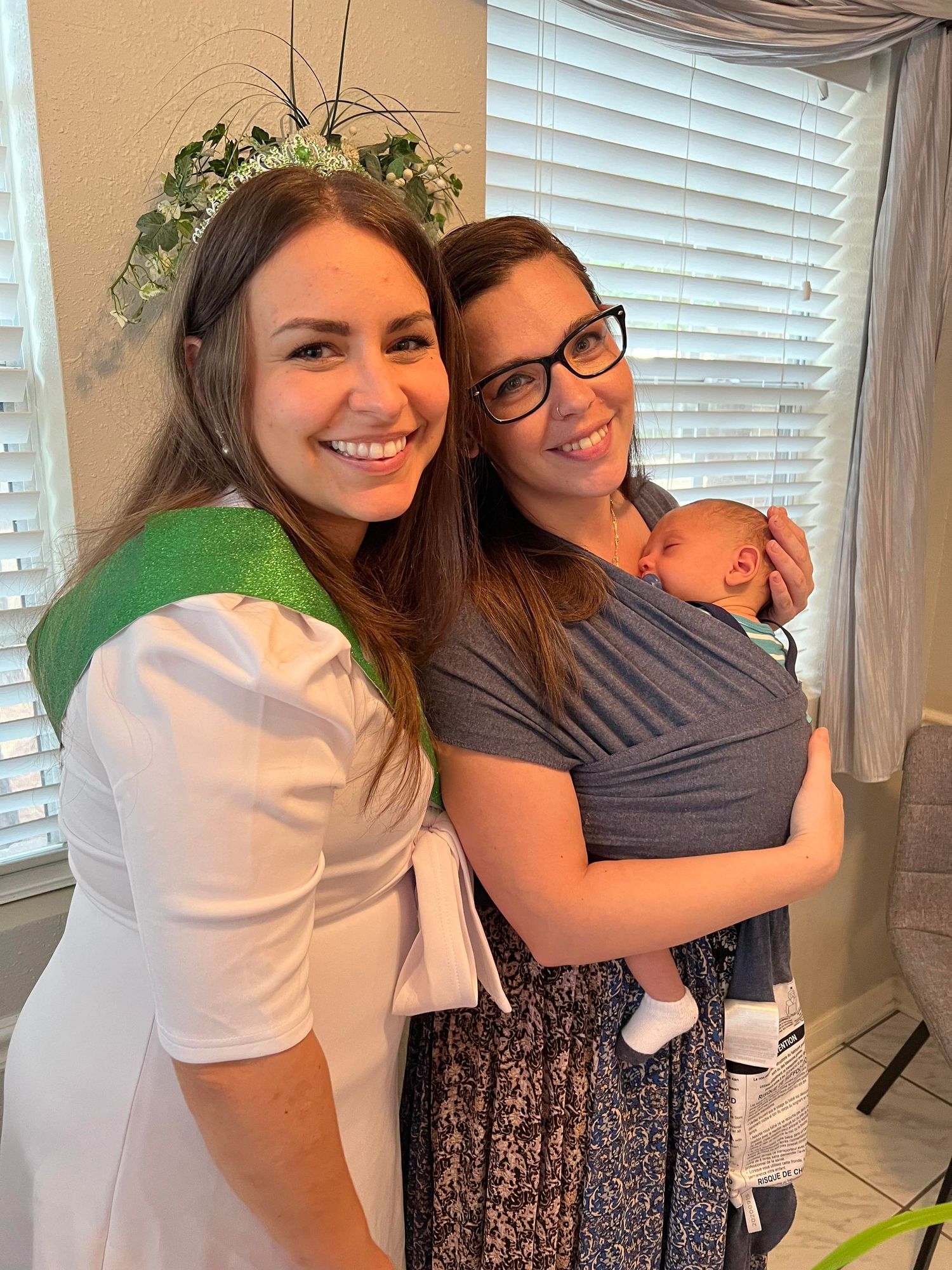 a brunette woman wearing a white dress and green sash poses next to another woman holding an infant