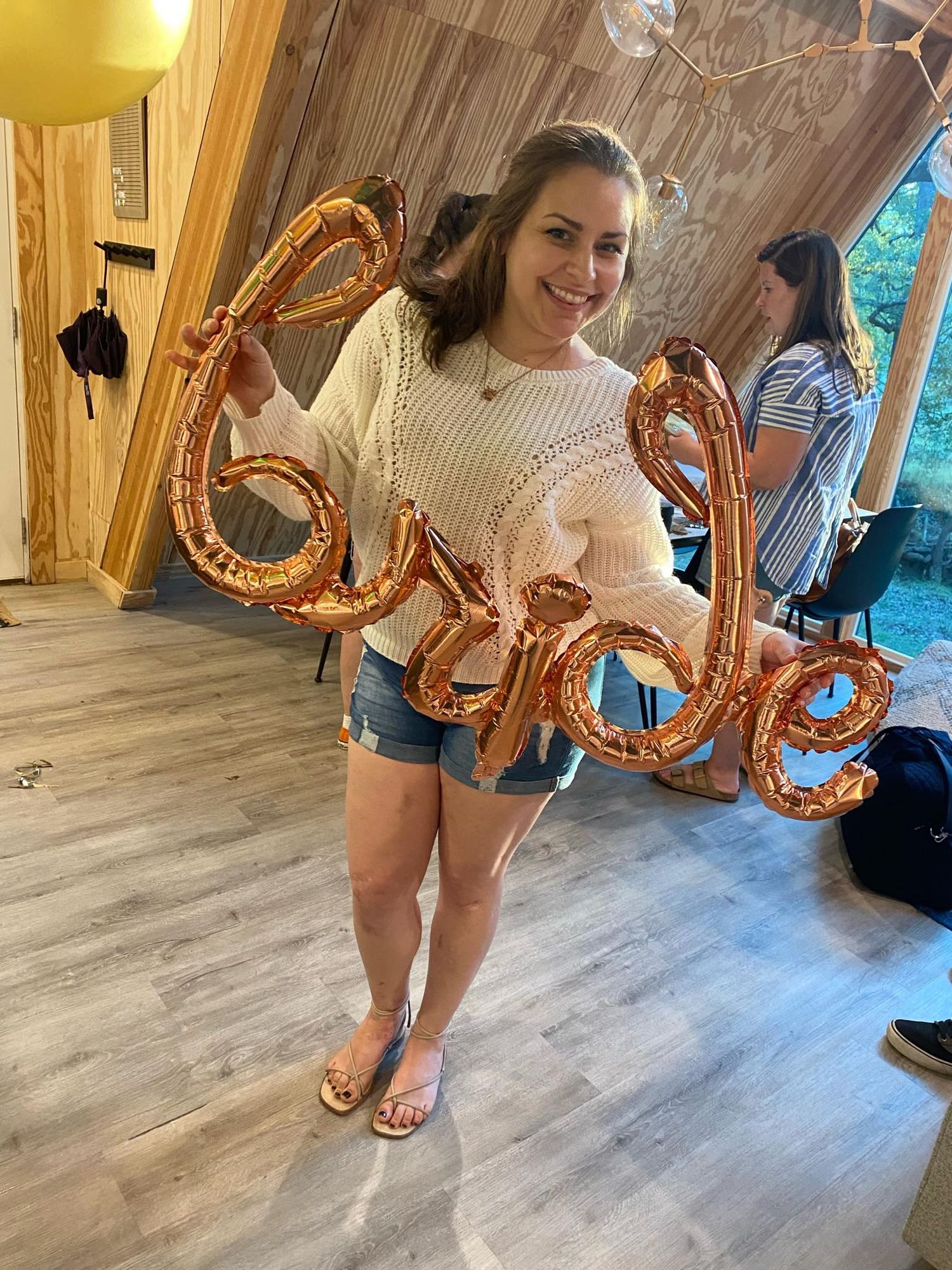 a brunette woman wearing a white sweater and denim shorts holds a rose gold balloon reading "bride"
