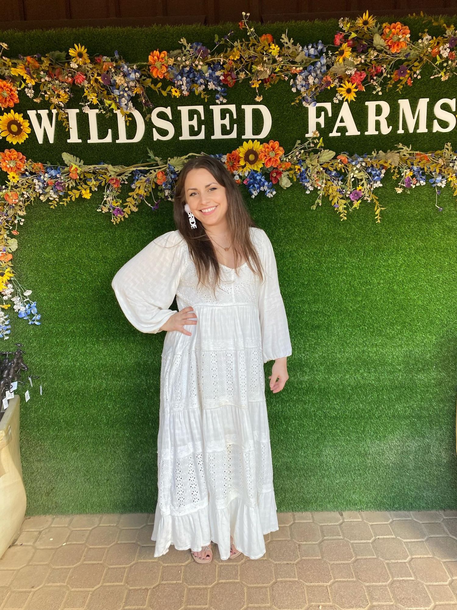 a brunette woman wearing a white dress poses with her hand on her hip in front of a grass backdrop