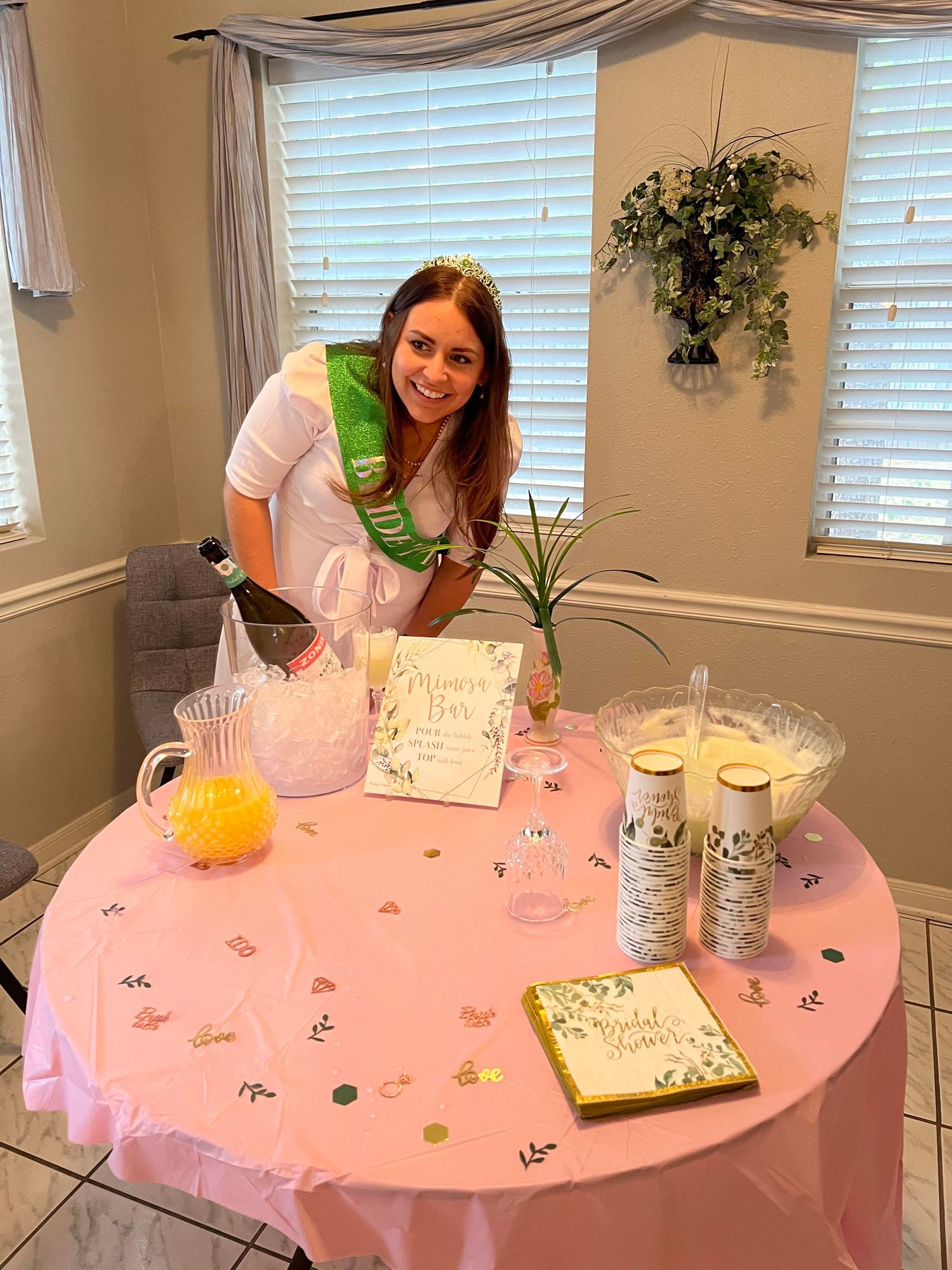 a brunette woman wearing a white dress and green sash bends down toward a pink table and smiles