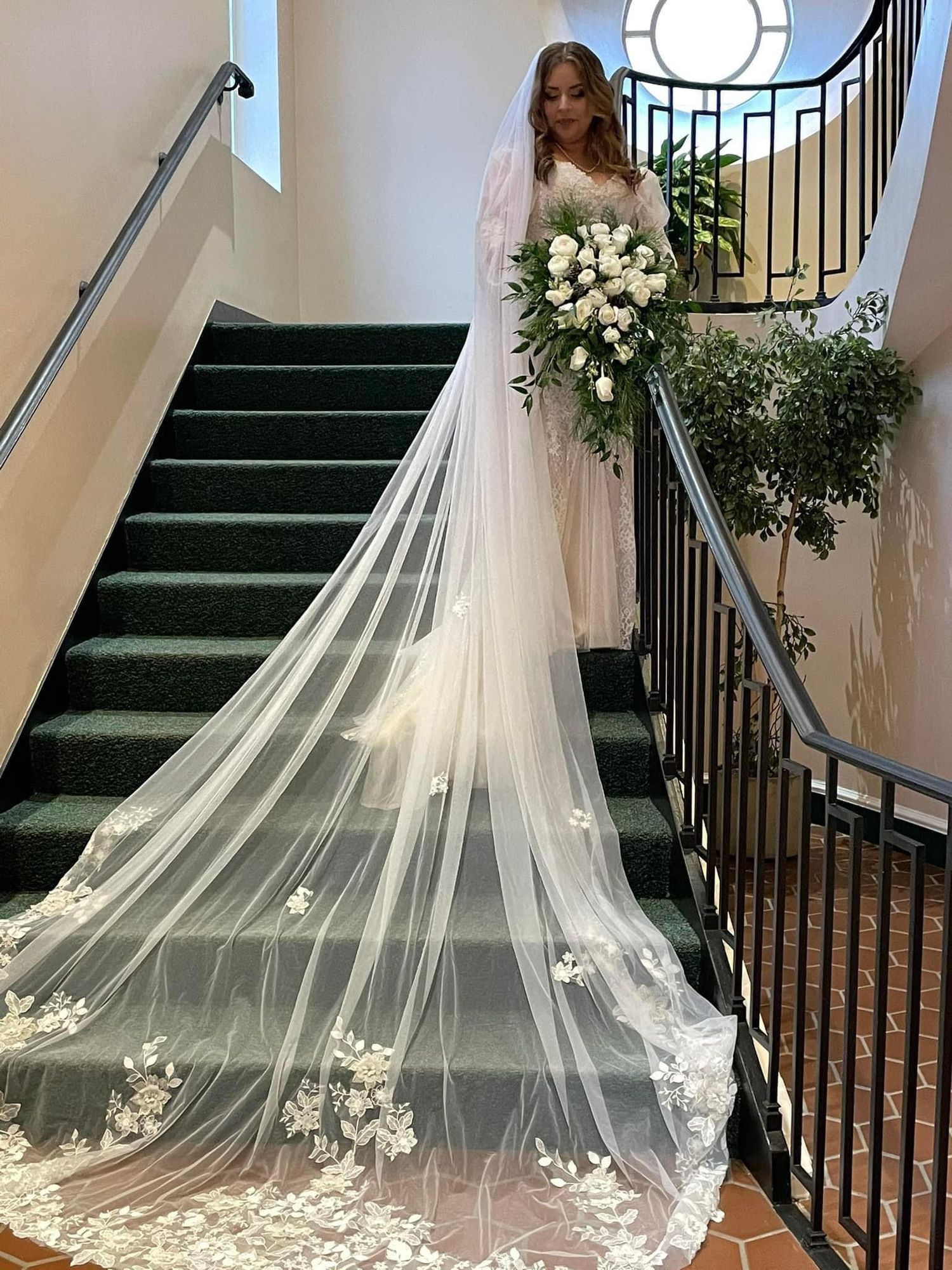 a bride wearing a white wedding dress stands at the top of green carpeted stairs with her veil stretched out