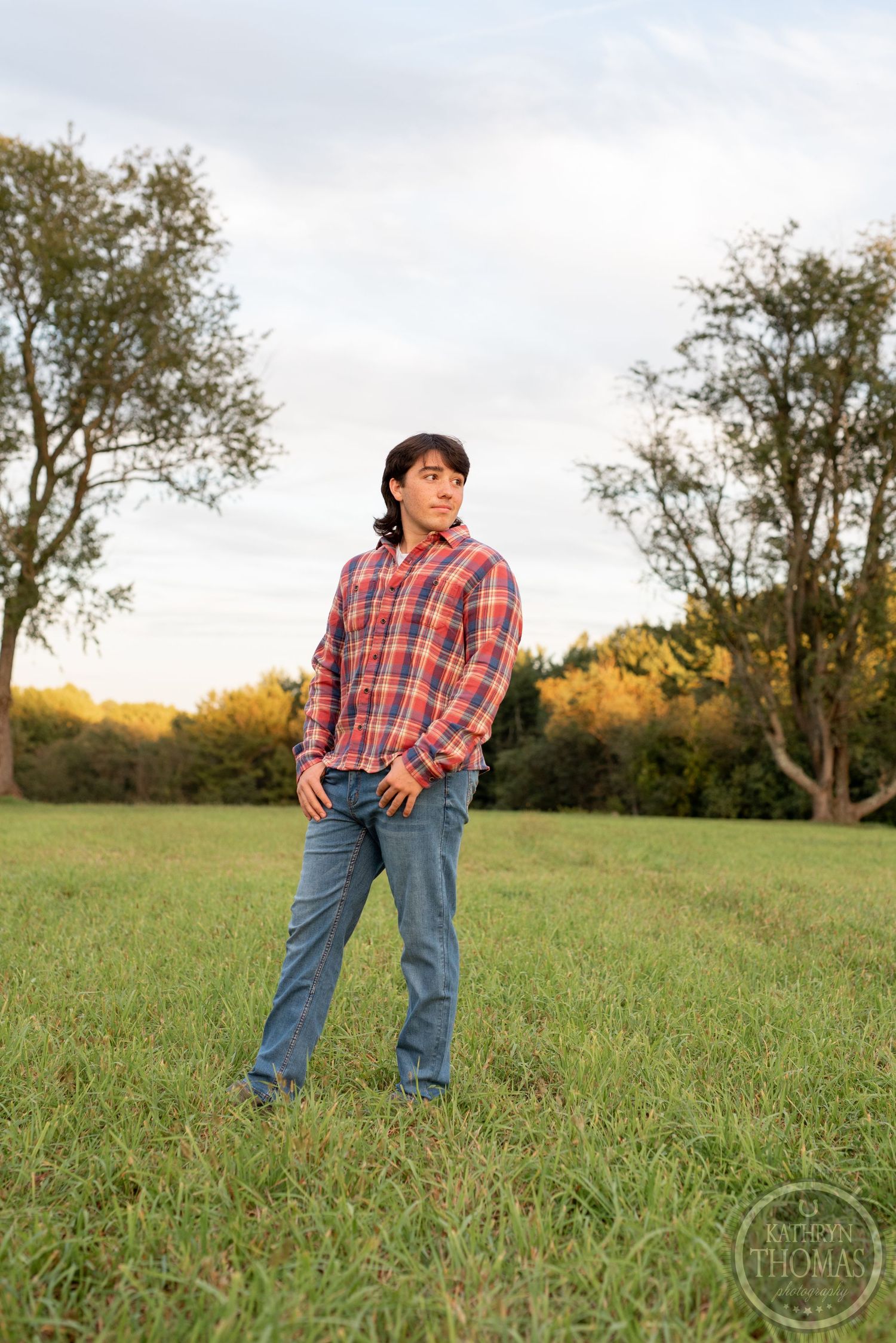 Person wearing plaid shirt and jeans poses casually in an open field during golden hour.