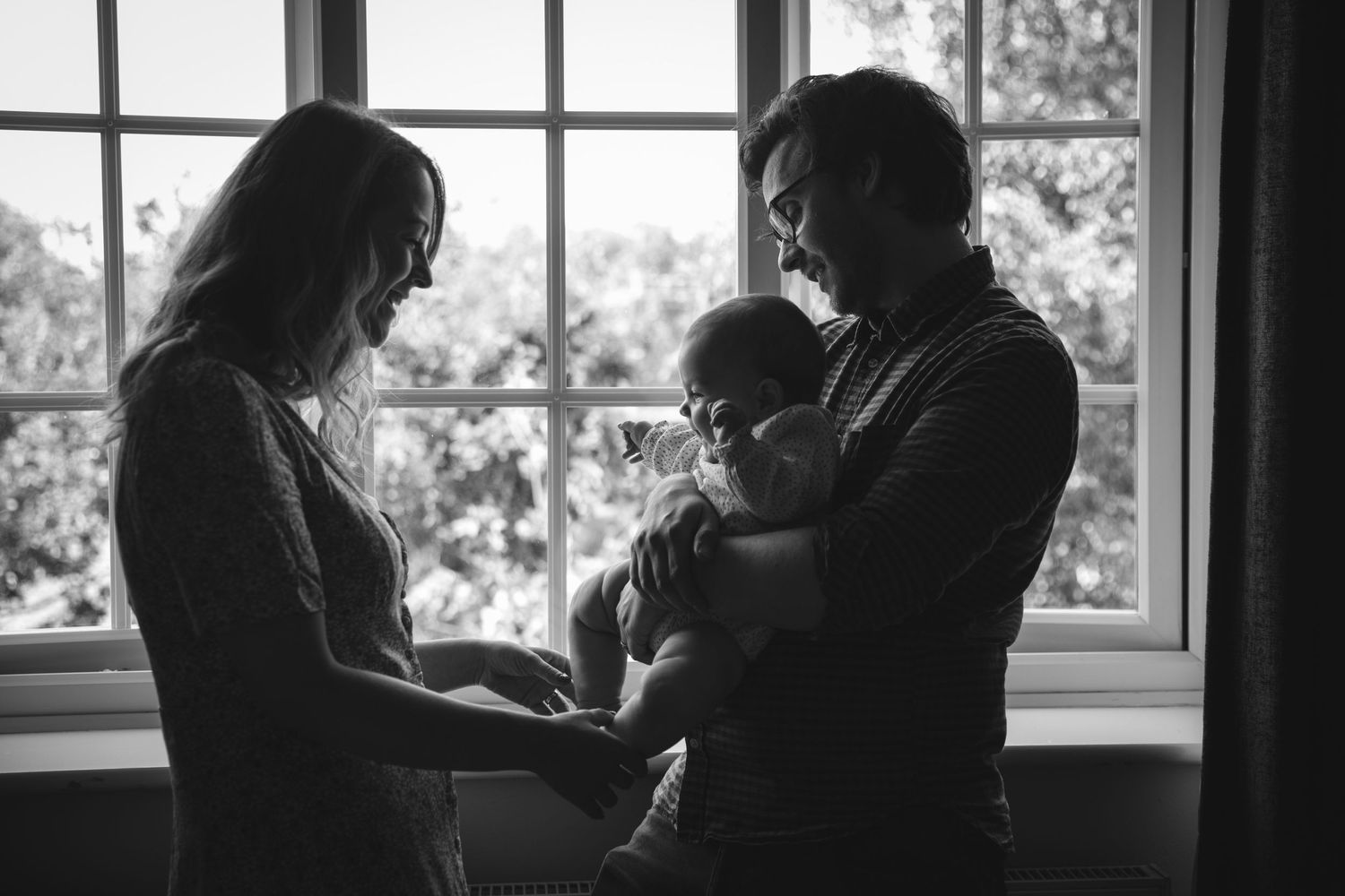 A tender black and white family portrait by a window with a baby.