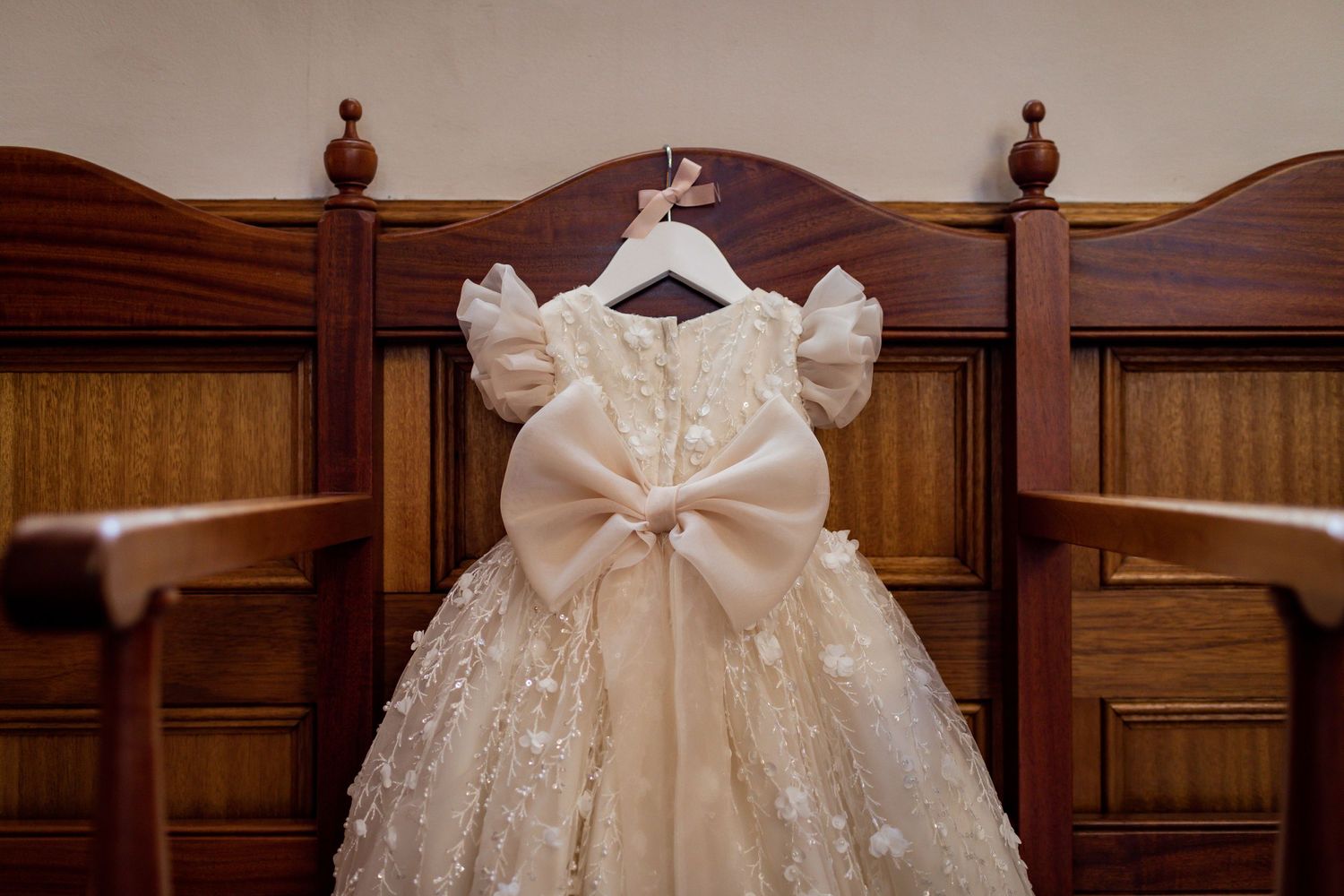 Close-up of an ornate white baptismal dress with bow detail hanging on wooden church pew.