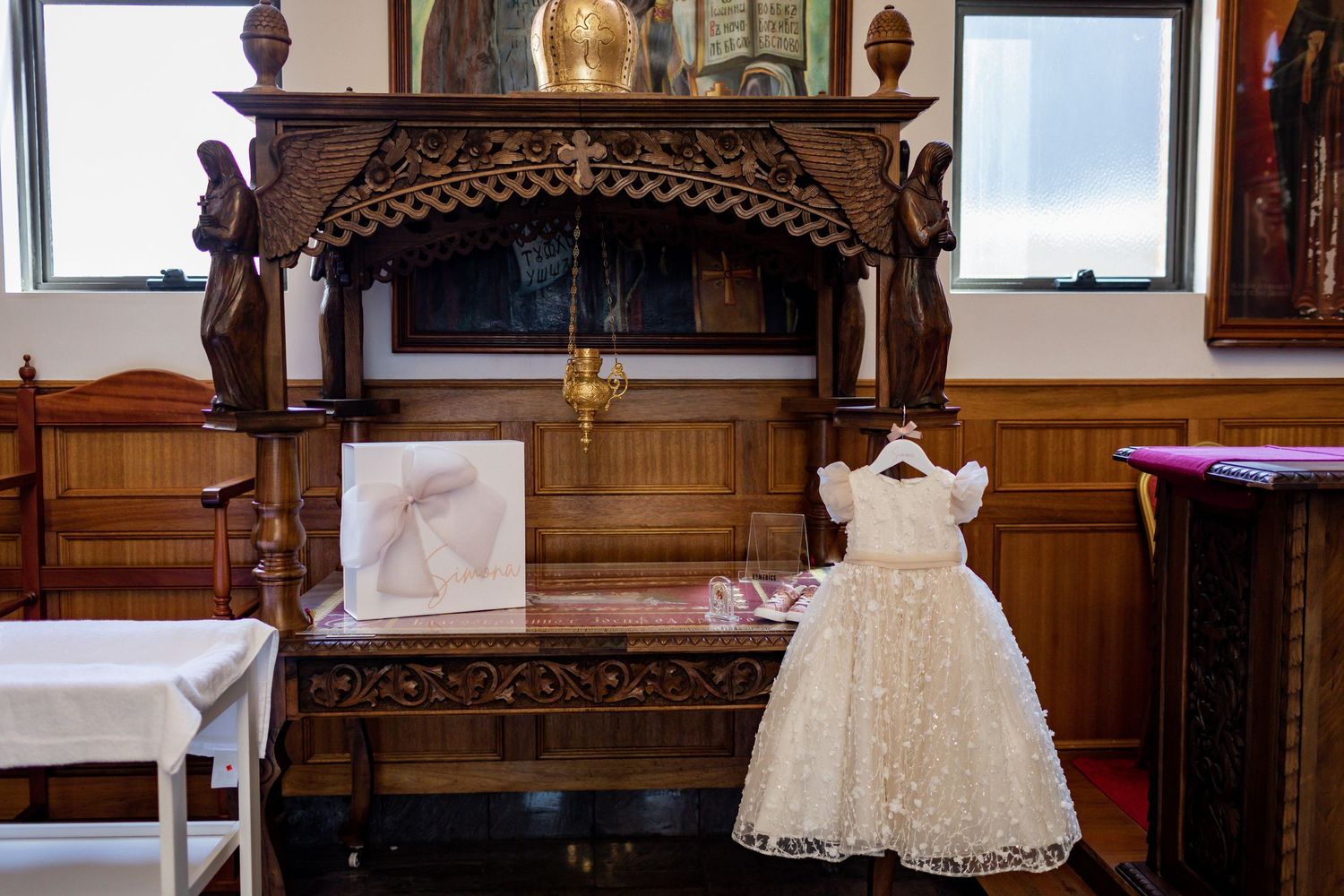 A delicate white lace baptismal dress displayed in an ornate wooden church setting with religious decor.