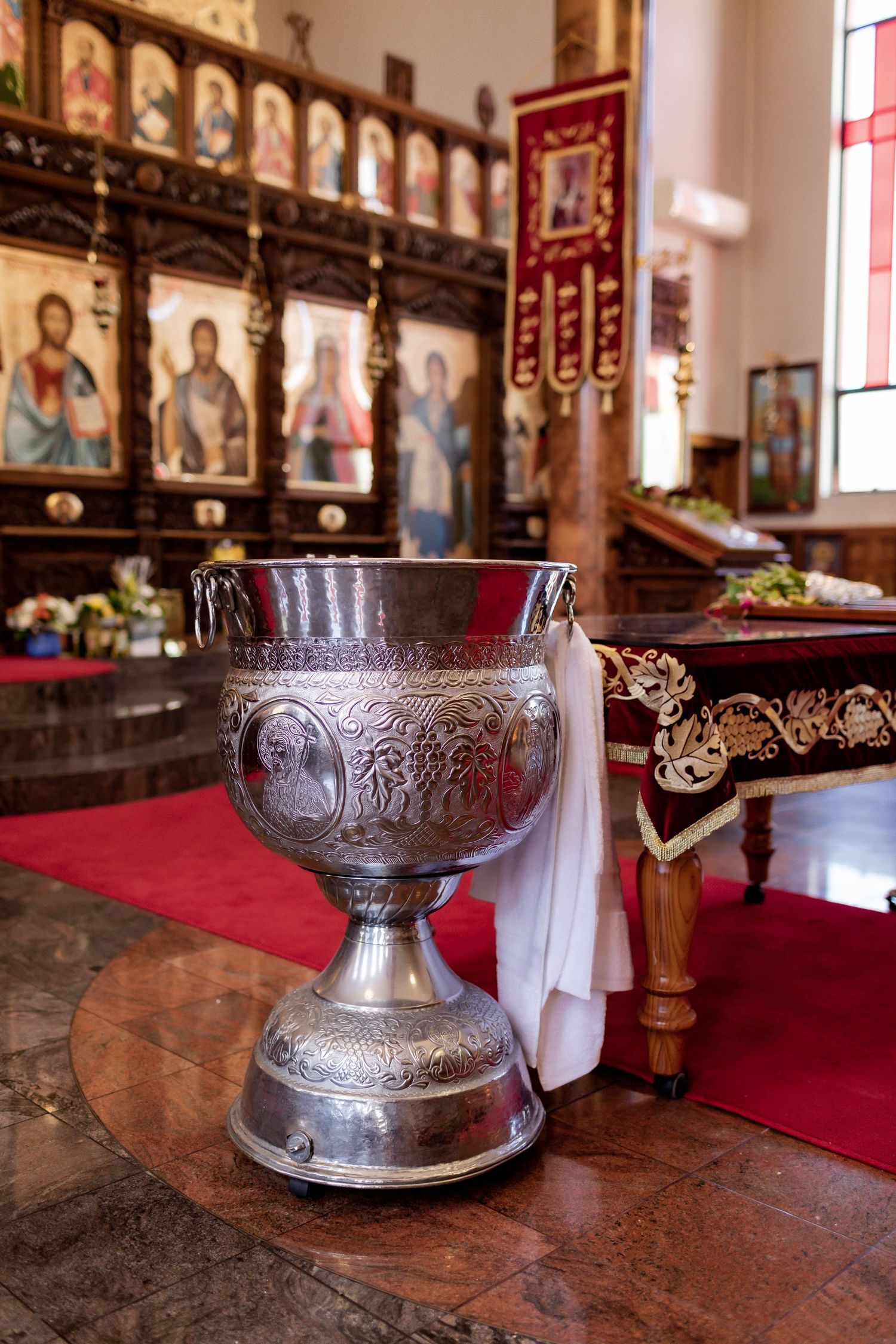 Ornate silver chalice displayed on red altar cloth with religious icons in background.